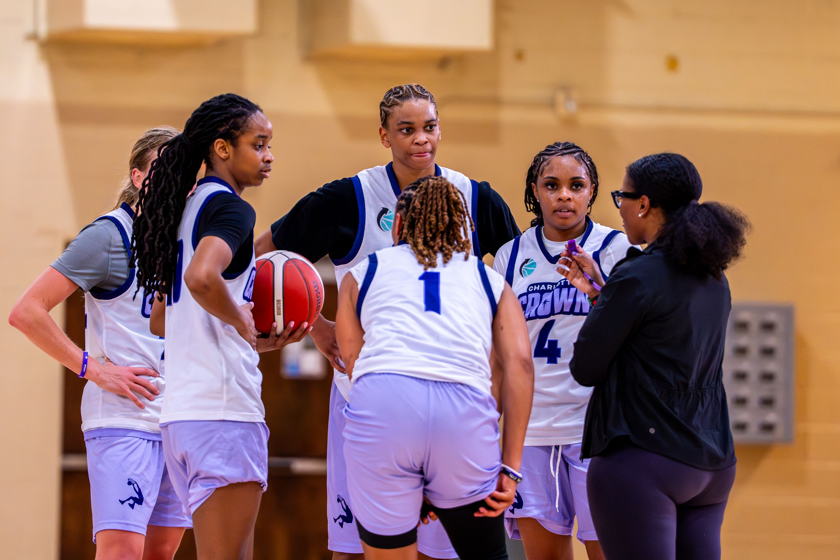 A group of female basketball players in white and blue uniforms huddle in a gym, listening to a coach. One player holds a basketball as they discuss strategy.