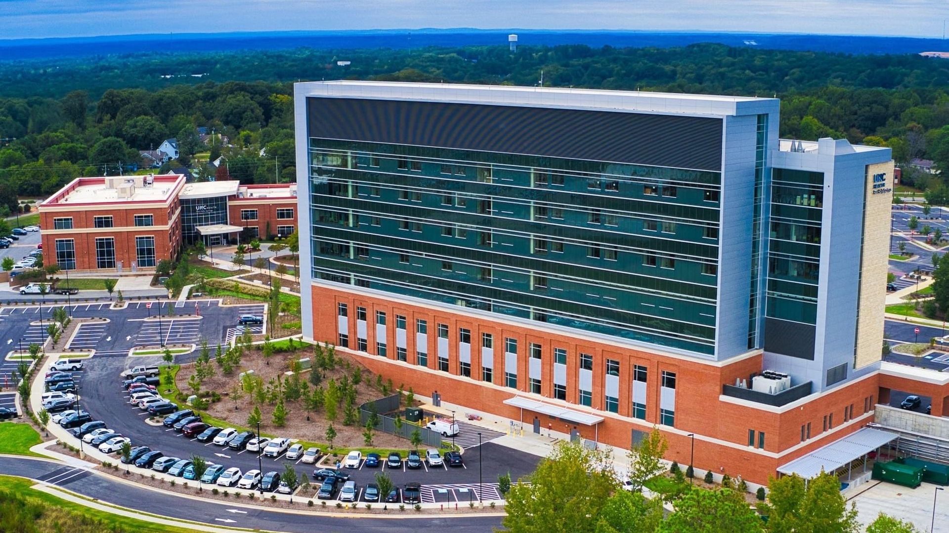 A high-rise hospital surrounded by a parking lot, trees and a pond.