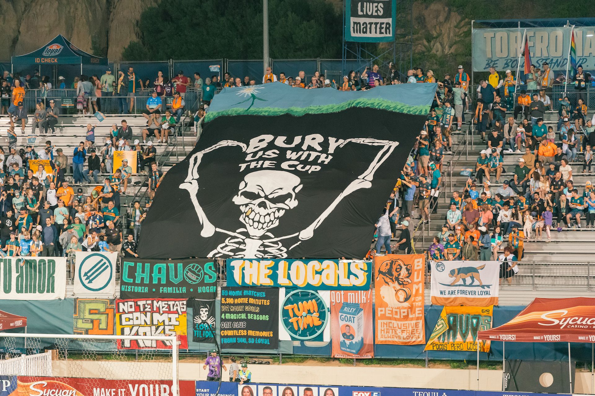 Fans in a soccer stadium hold up a giant banner that reads "Bury us with the cup" with a skeleton. 