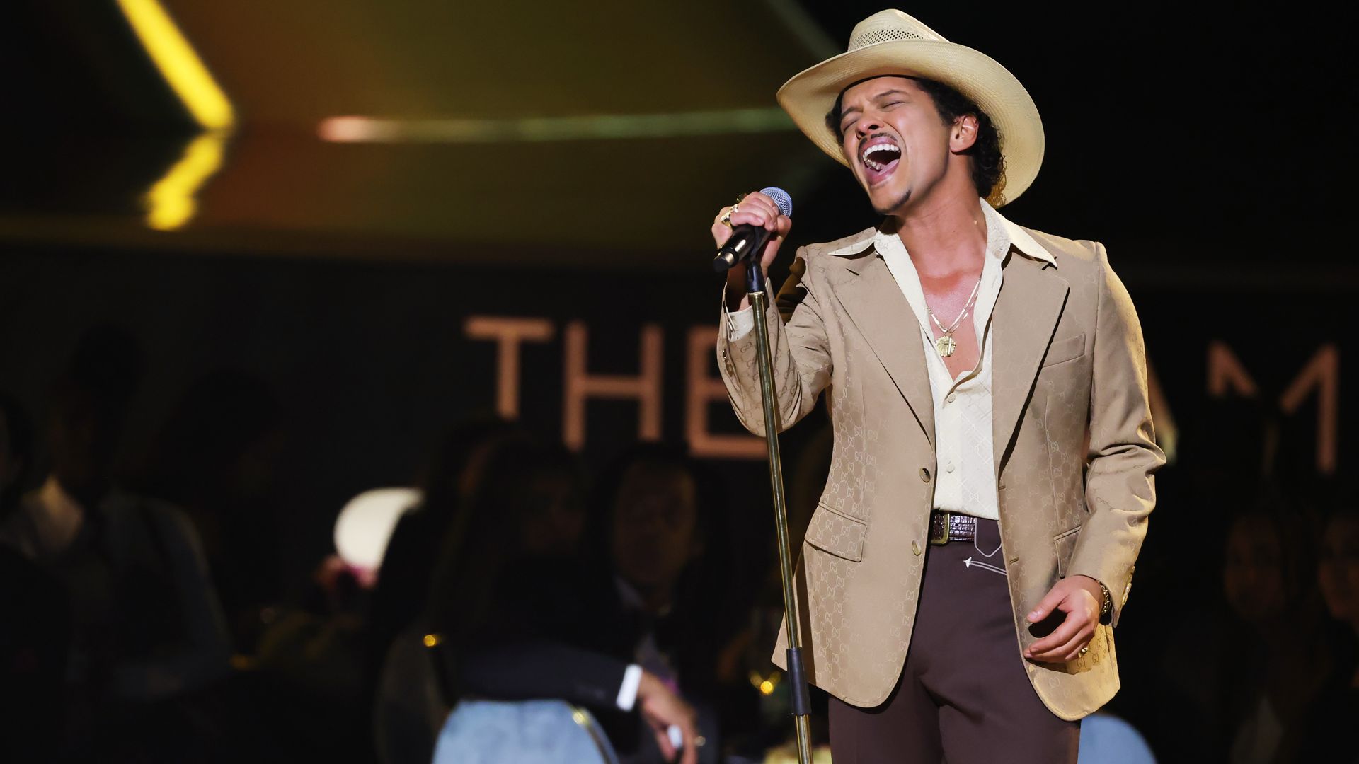 Bruno Mars, wearing a beige jacket, white shirt, brown pants, and cream cowboy hat, passionately sings into a microphone at the 2025 Grammy Awards.