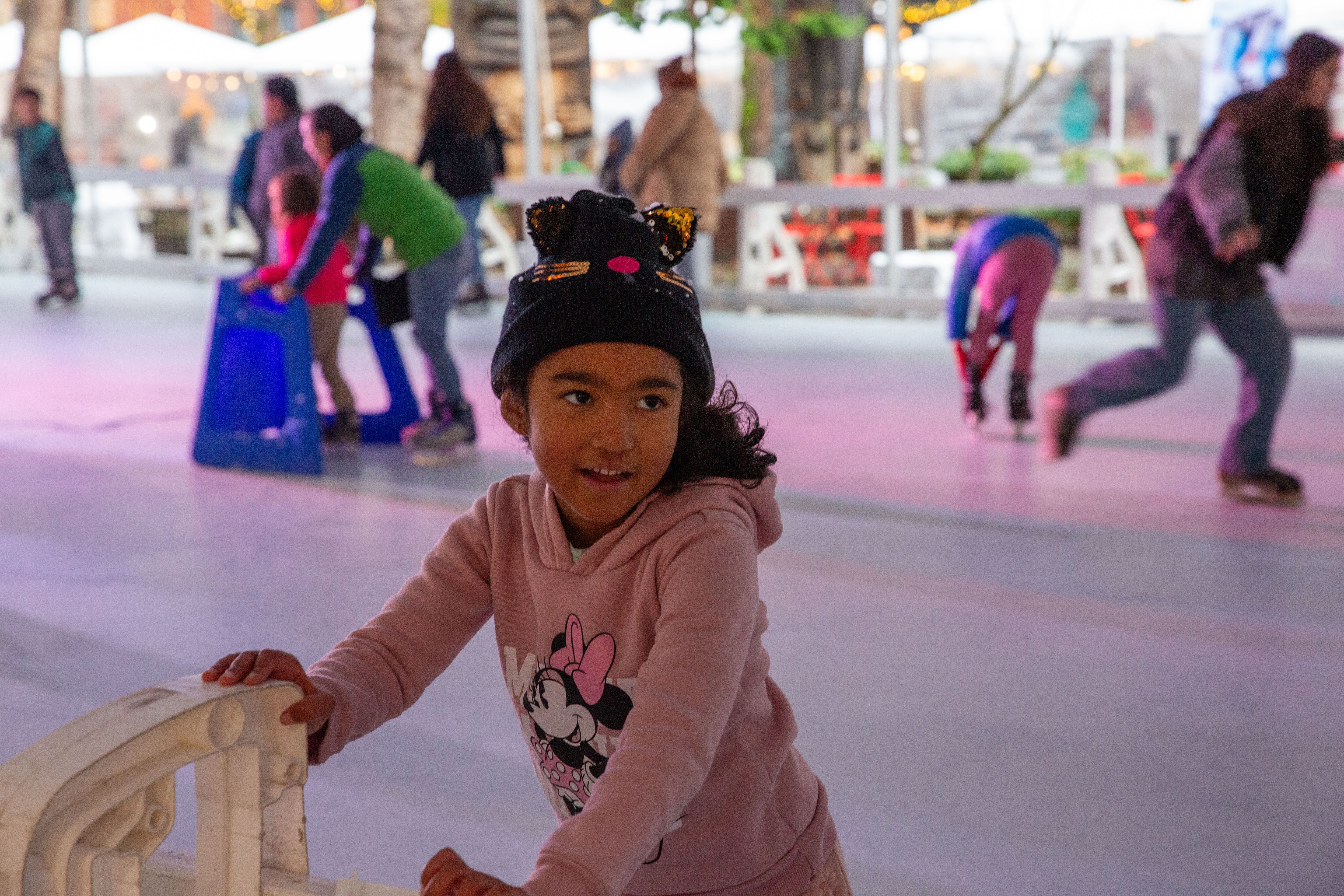 A girl with a knit hat in a pink Mickey Mouse sweatshirt stops and looks over her shoulder on an ice skating rink. 