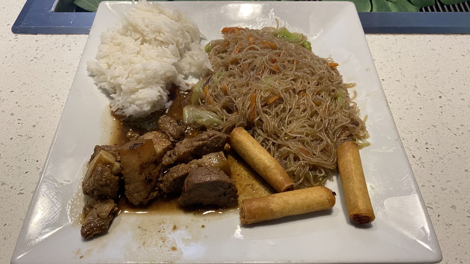 A square plate with white rice, thin noodles, egg rolls and chunks of pork in brown sauce. 