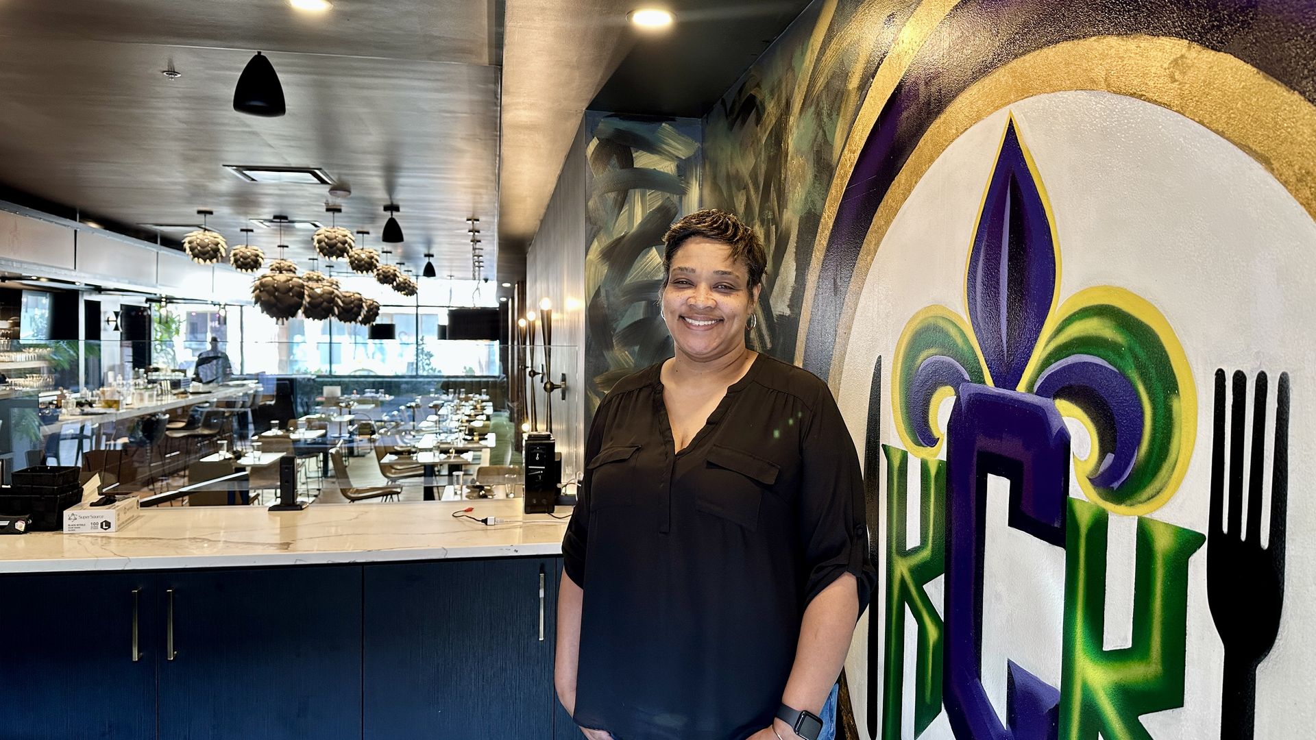 Smiling woman in a black blouse stands beside a colorful mural featuring a fleur-de-lis and utensils, inside a modern restaurant with blue cabinetry, marble counter, and a bright dining area.