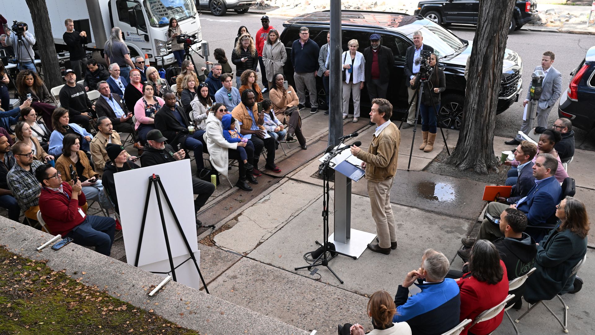Outdoor press briefing: a man speaks from a podium to a diverse crowd seated on folding chairs; photographers film as cars and a black SUV line the street behind them.
