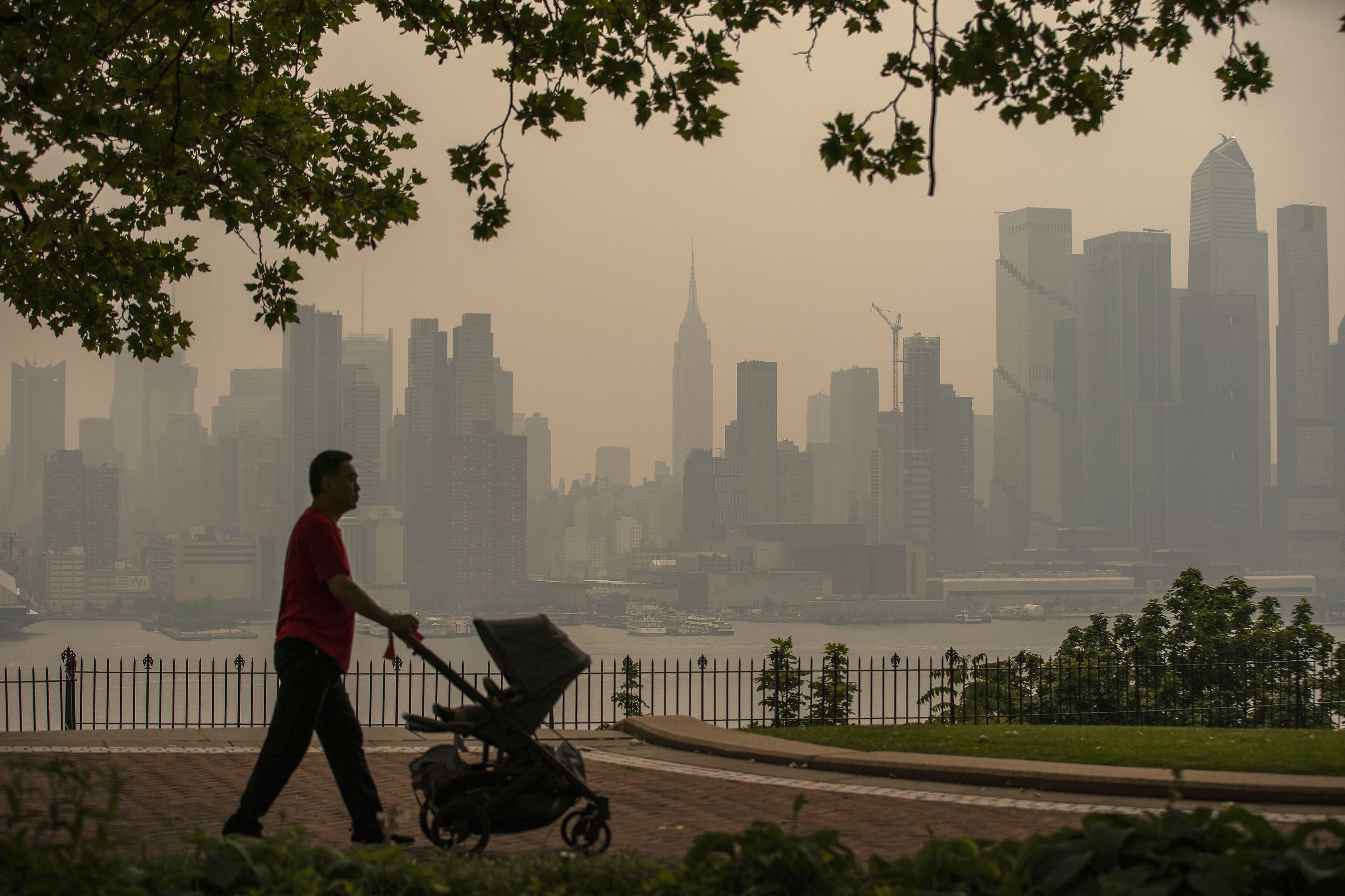 Man with baby stroller walks with Manhattan skyline in background.