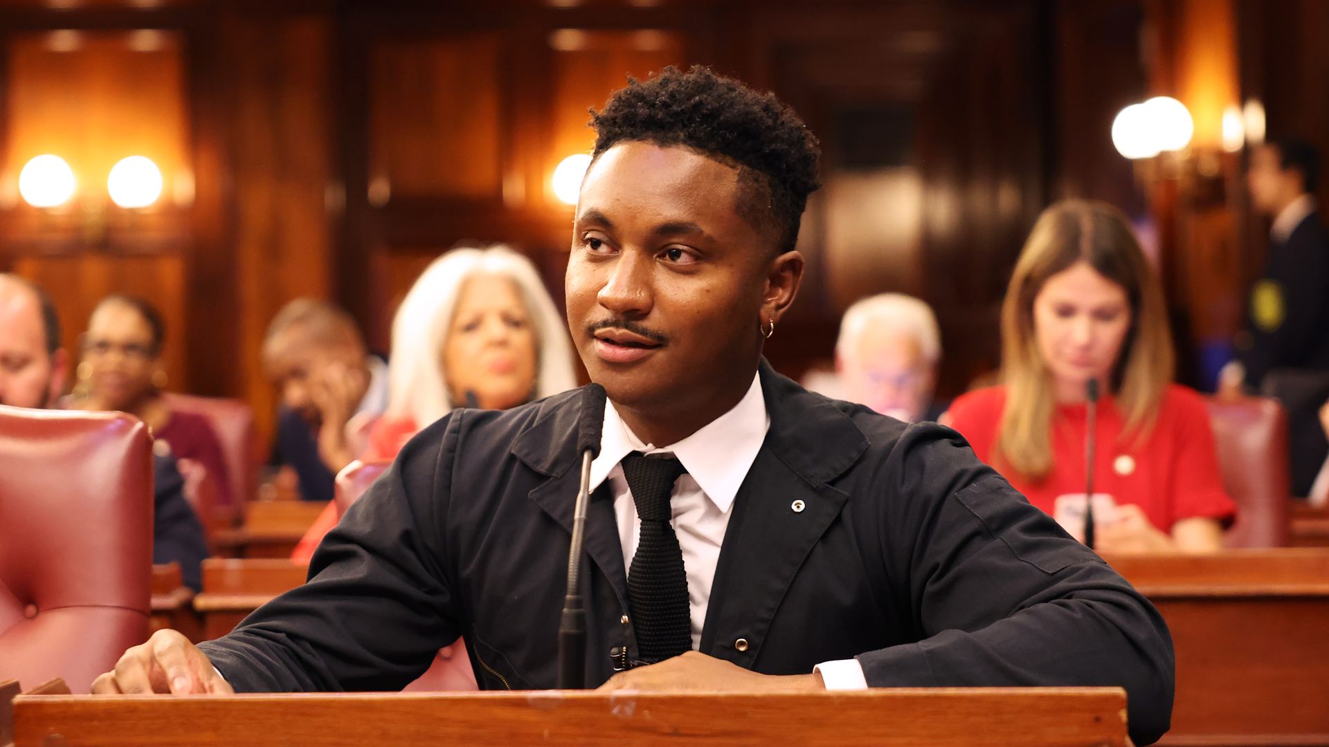 New York City Council member Chi Ossé, dressed in a black jacket, white shirt and black tie, sits at his desk in council chambers.