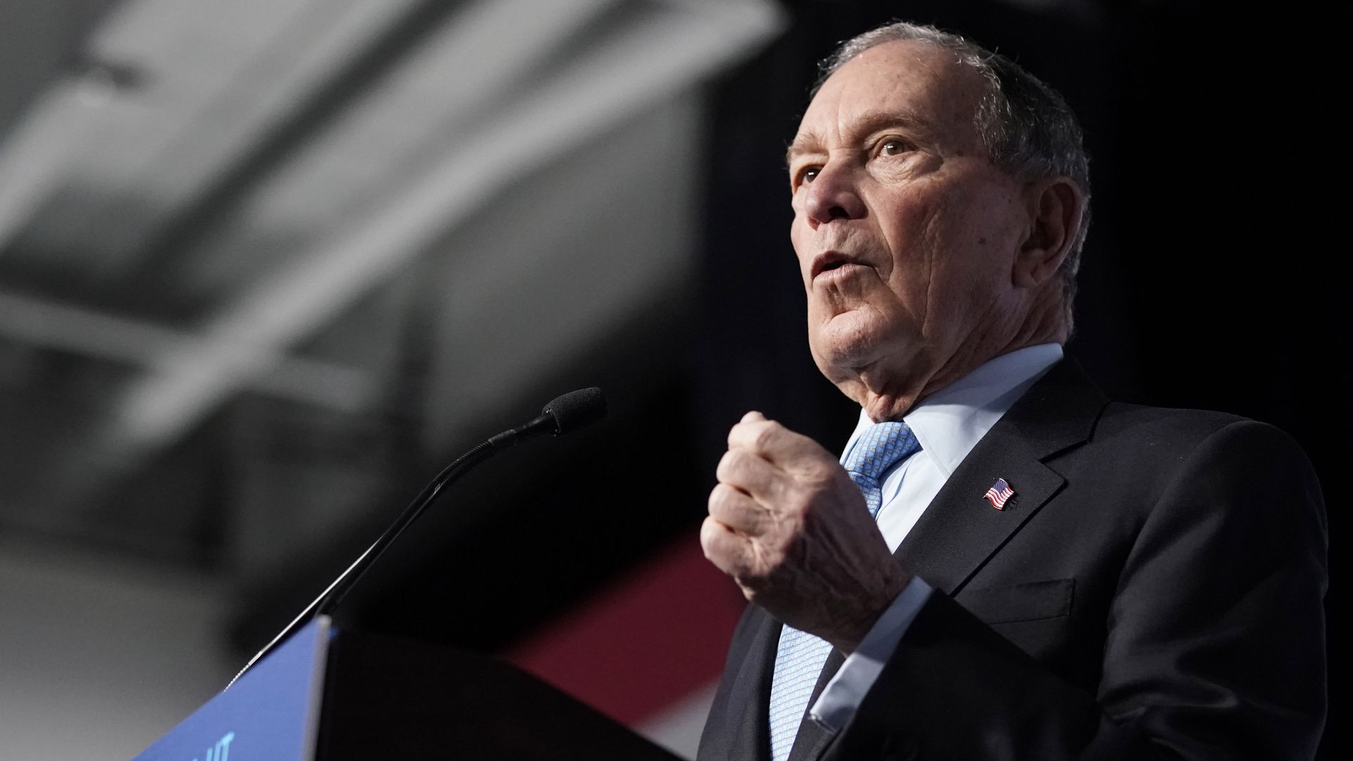 In this image, Michael Bloomberg stands at a podium in a suit.