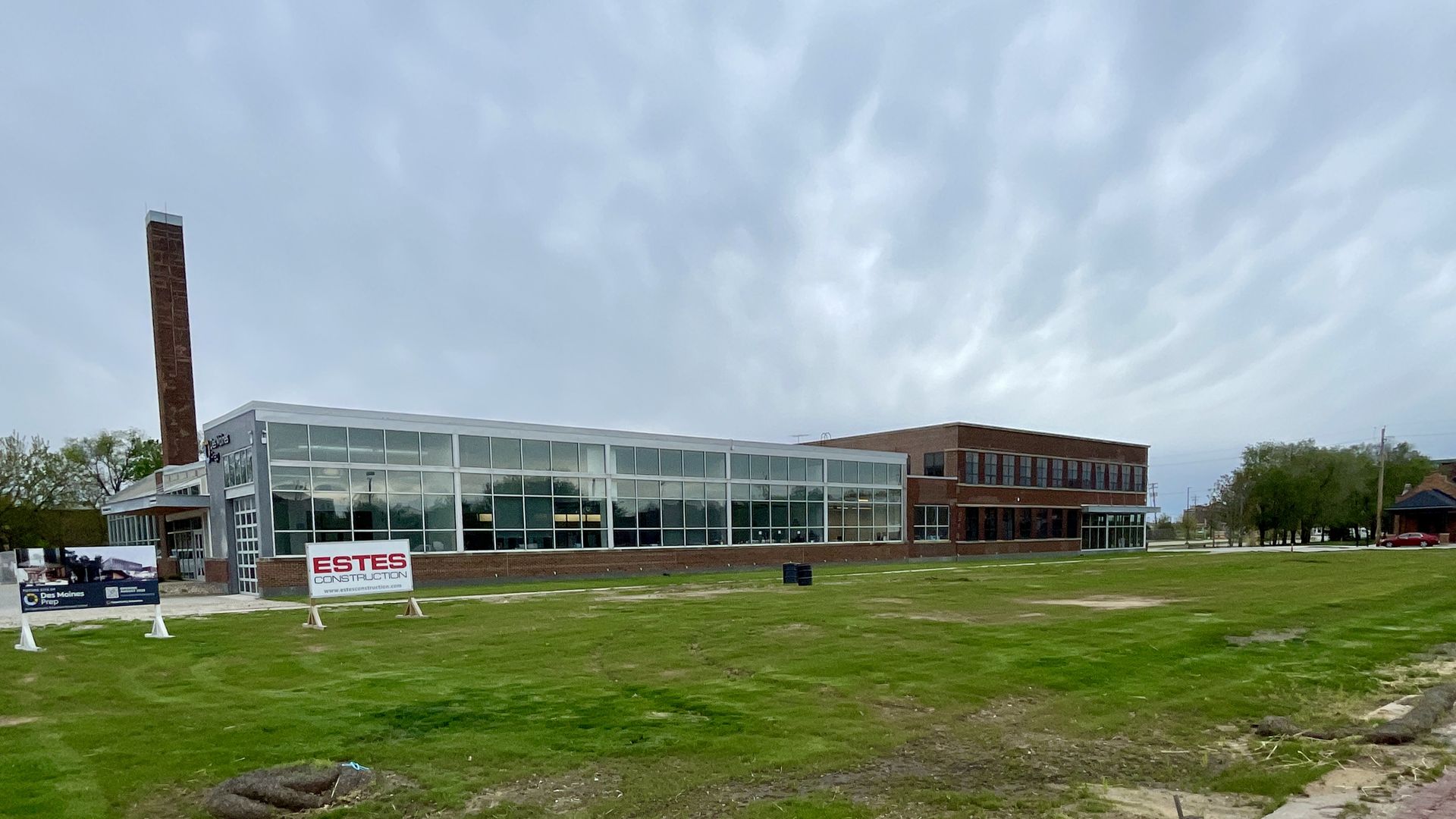 Modern brick-and-glass office building with a tall brick smokestack on the left, large front windows, and construction signs on a grassy lot under a cloudy blue sky.