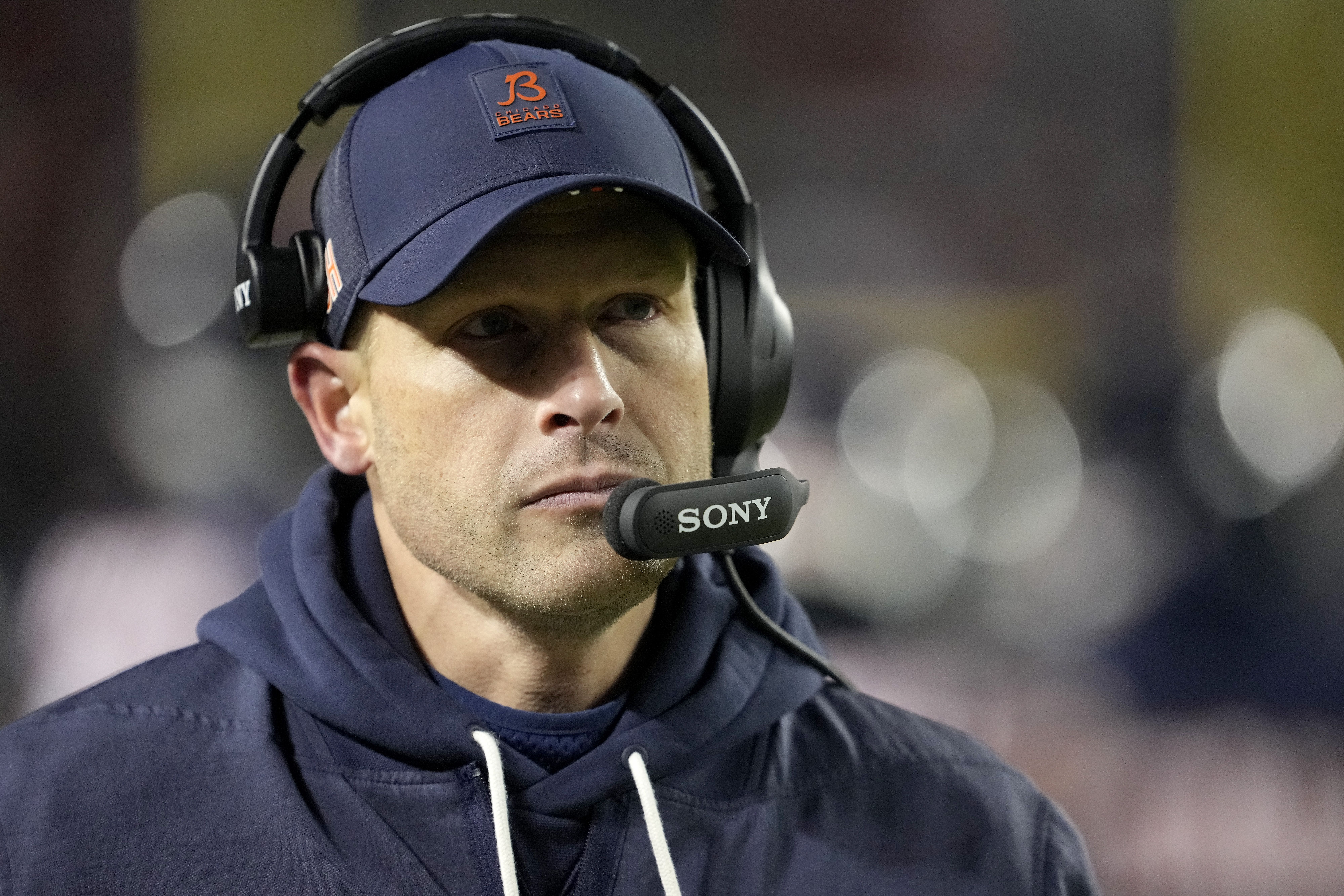 Man wearing a navy Chicago Bears cap and hoodie with a black Sony headset microphone, focused expression, blurred background with lights.
