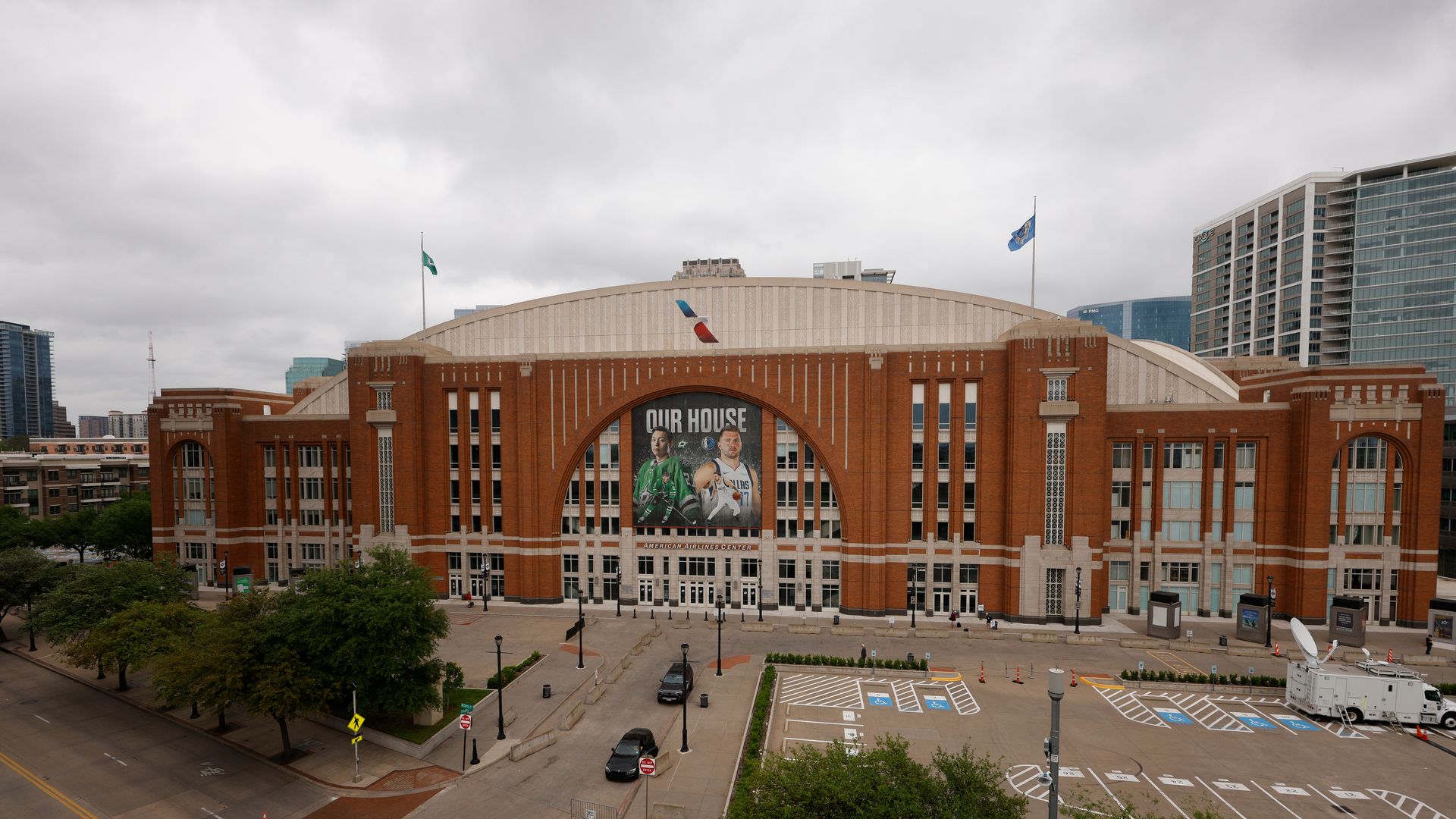 An external view of American Airlines Center with brown brick facade, large arched windows, and a banner featuring athletes with text "OUR HOUSE" under cloudy sky.