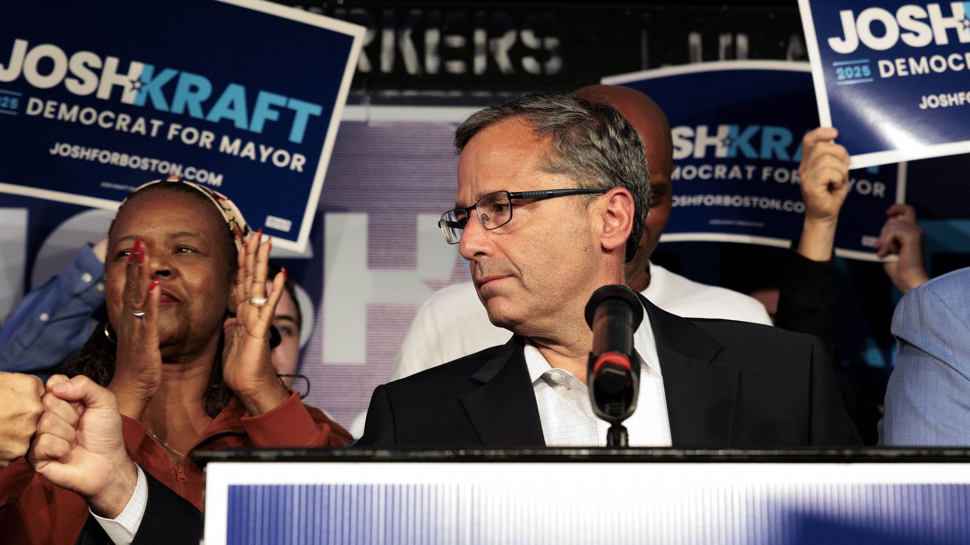 Josh Kraft looks to his right and fist bumps someone out of view while delivering remarks at an election night watch party for his mayoral campaign. He's surrounded by supporters holding up his signs while he's at the podium. 