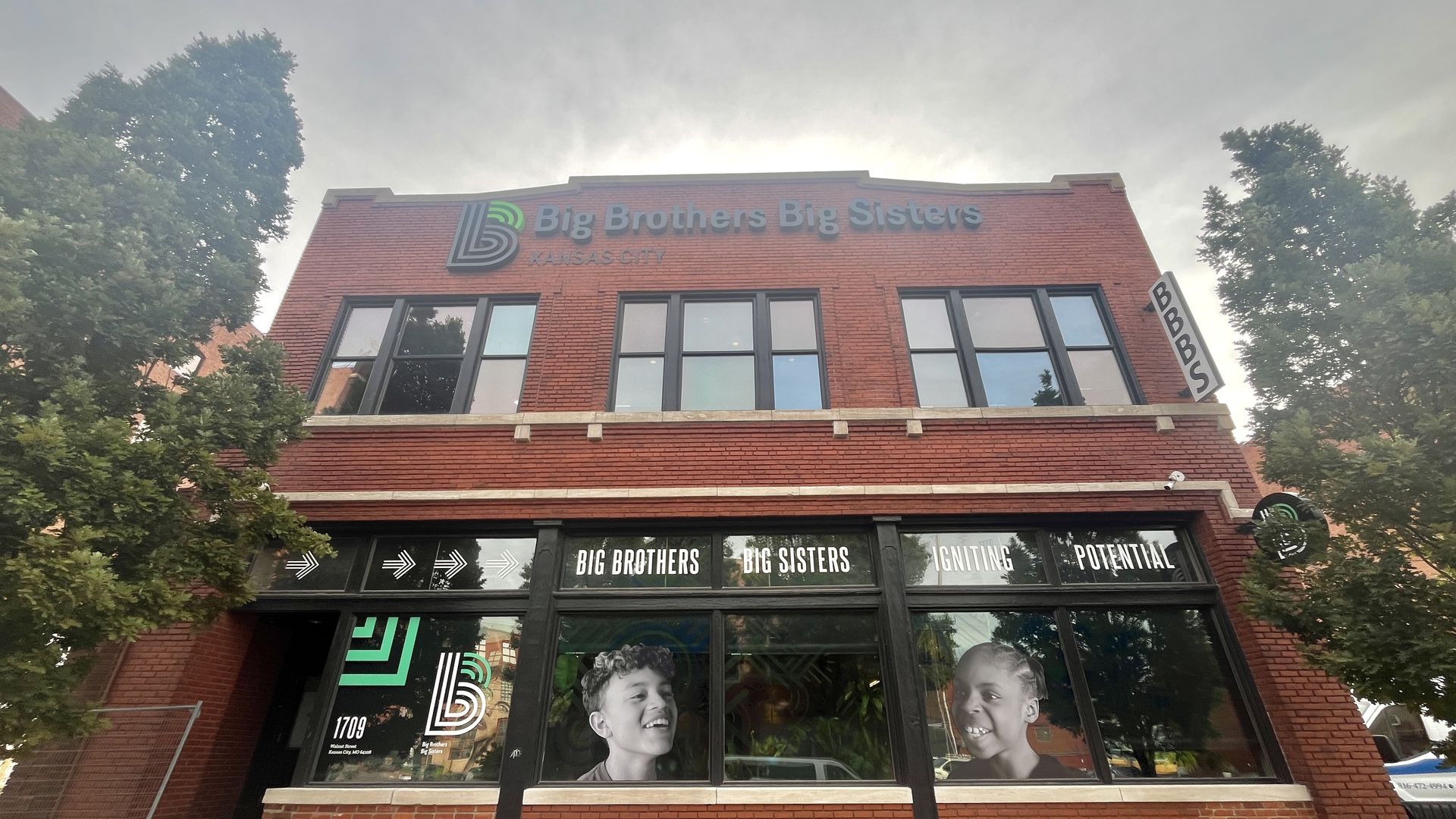 Red brick building with Big Brothers Big Sisters Kansas City signage, large windows featuring smiling children's portraits, and trees on either side under a cloudy sky.