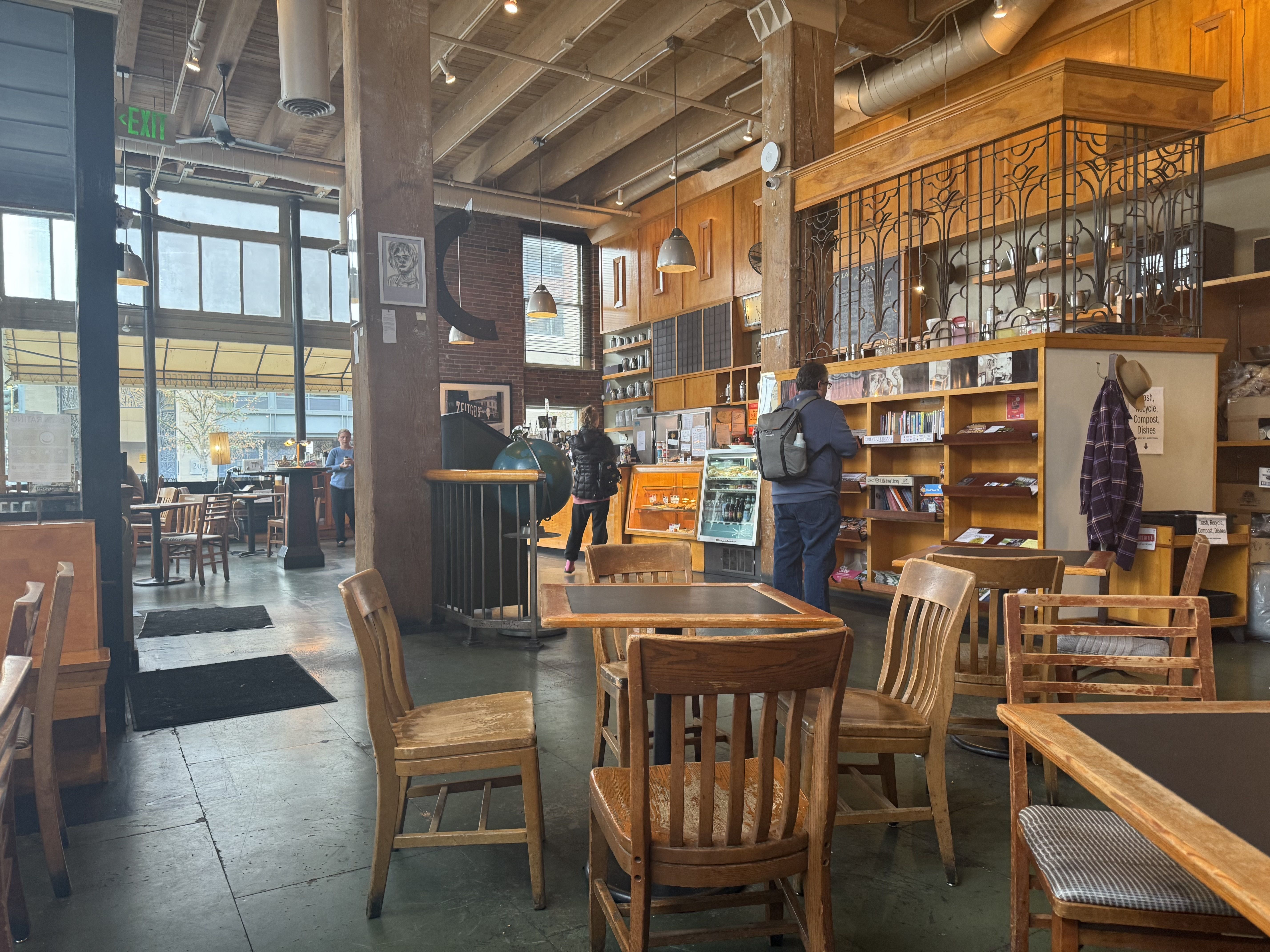 Interior of a cozy cafe with wooden tables and chairs, exposed brick, large windows, and warm lighting. Two customers near the counter and bookshelves add a welcoming touch.