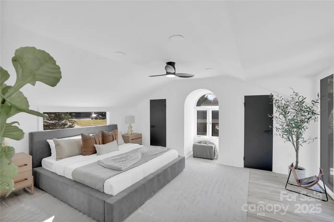 Bright bedroom with white walls, gray upholstered bed with brown and beige pillows, wooden nightstands, black doors, a ceiling fan, large green plant, and an arched window alcove with a gray ottoman.