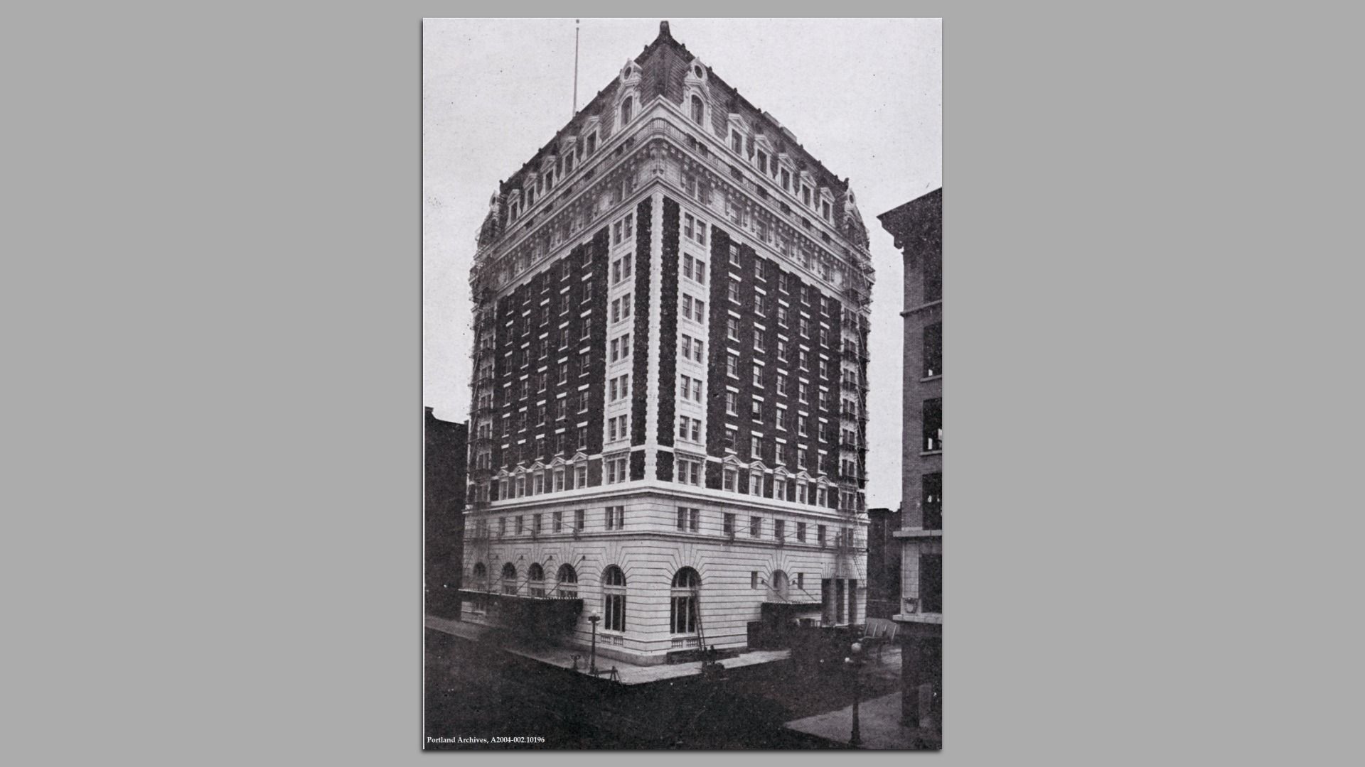 Black and white photo of a tall, ornate brick and stone building with arched windows at the base and elaborate rooftop architecture, viewed from street corner in an urban setting.