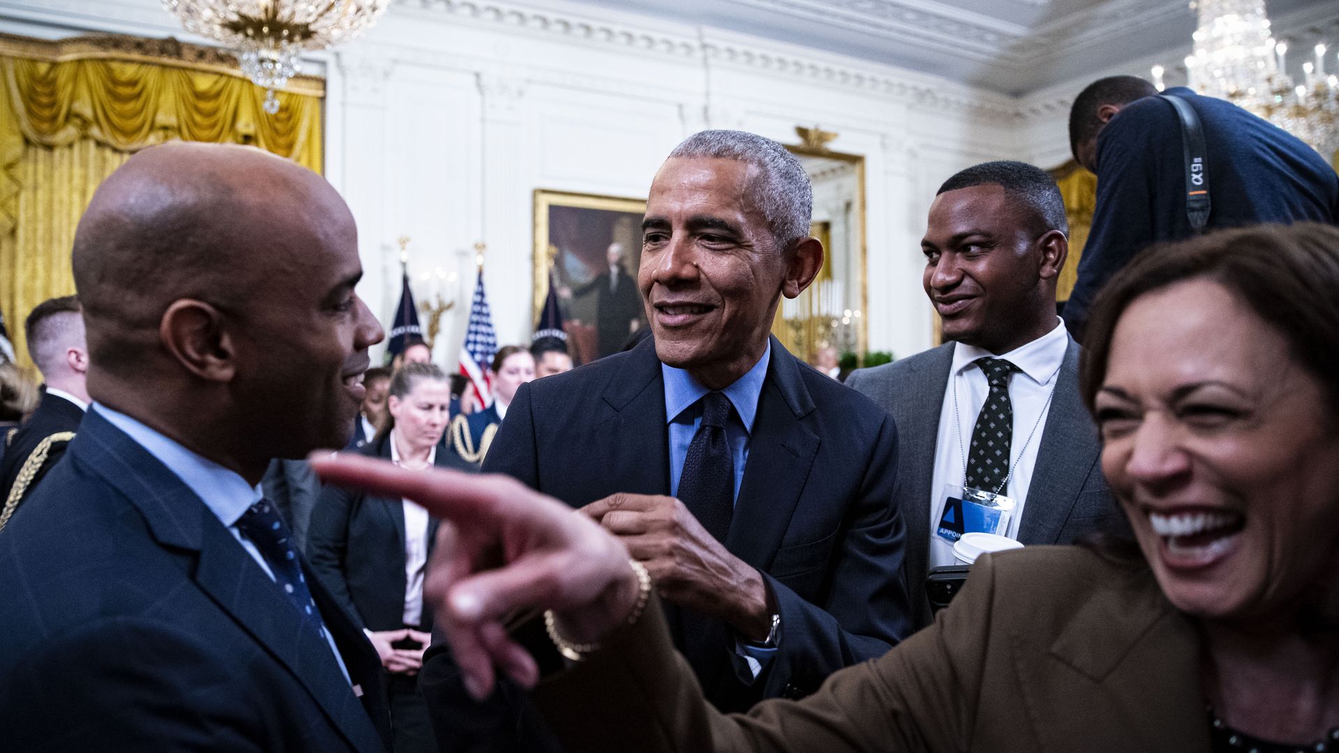 Former President Barack Obama, wearing a navy suit, shirt and tie, greets attendees at a White House event along with Vice President Kamala Harris.