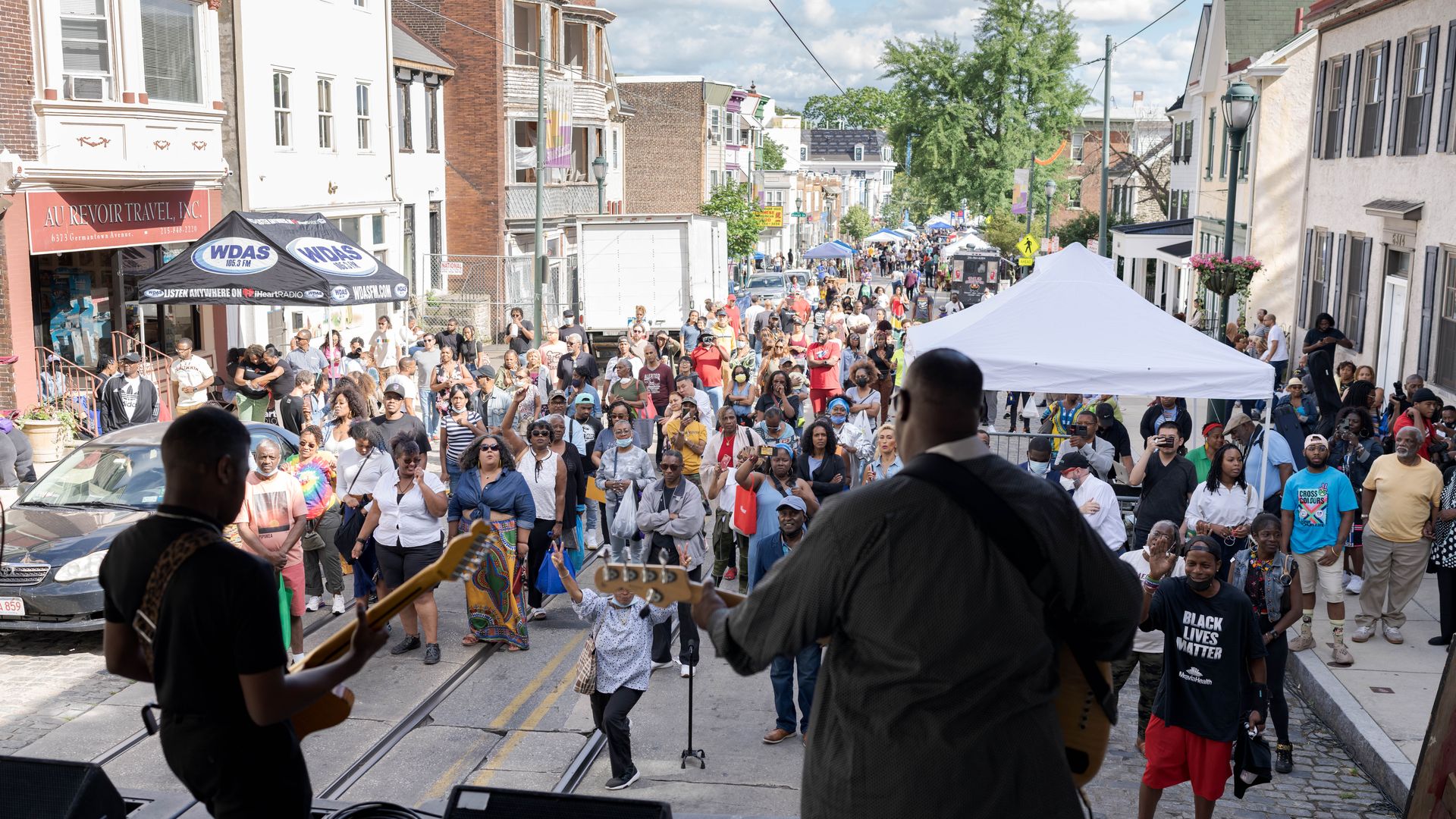 Crowd fills a city street for a festival/parade, viewed from a musician on stage. People walk along the road among tents, storefronts, and colorful banners with trees and blue sky in the distance.