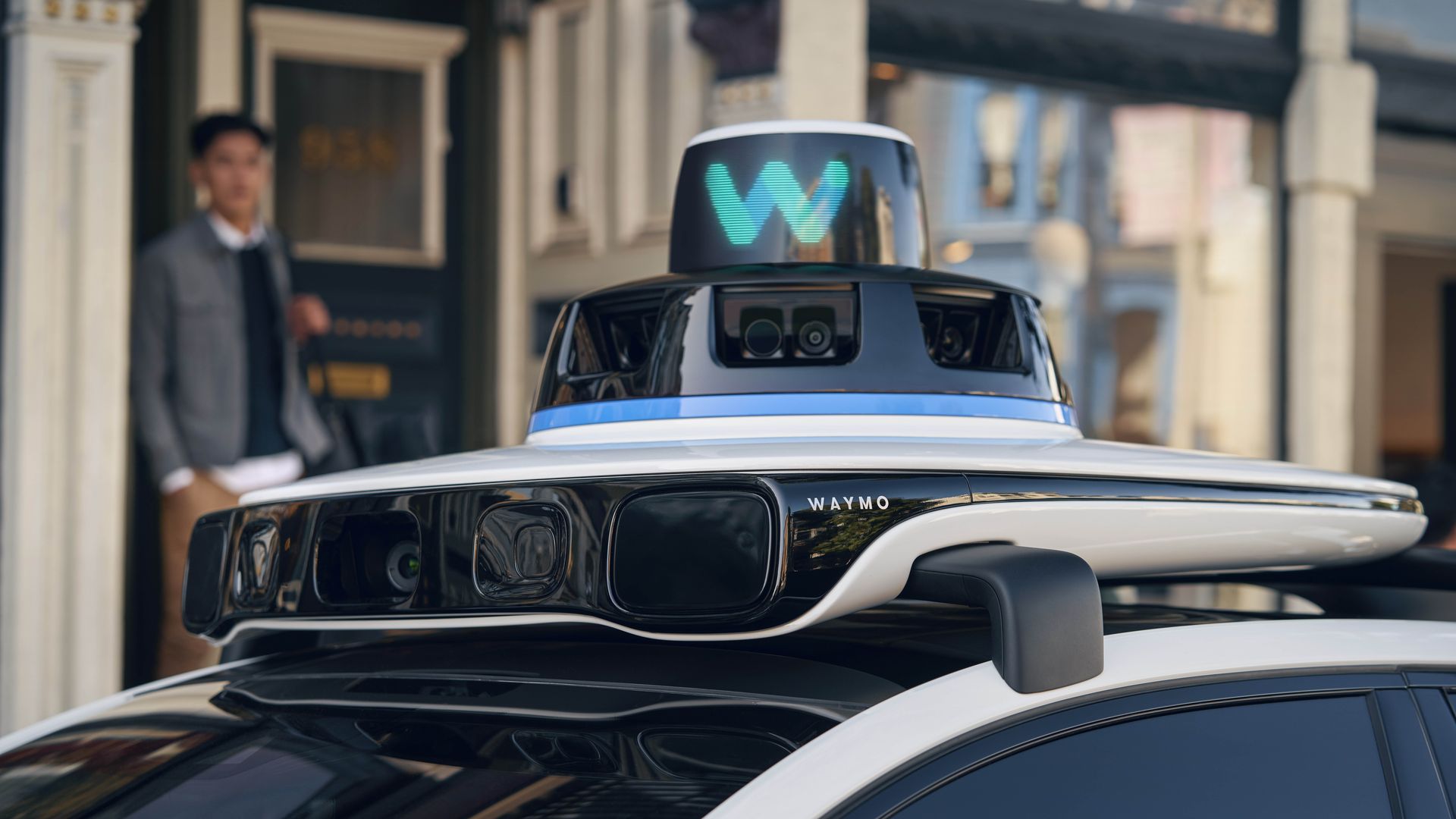 Close-up of a white Waymo autonomous vehicle's sensor array on the roof, featuring cameras and lidar with a digital blue "W" logo, with a young man in the background