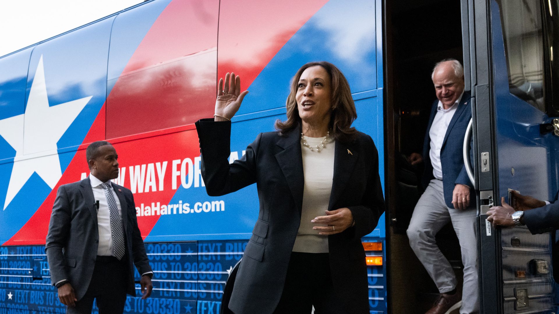 Kamala Harris waves in front of her campaign bus as Minnesota Gov. Tim Walz steps down the bus' stairs.