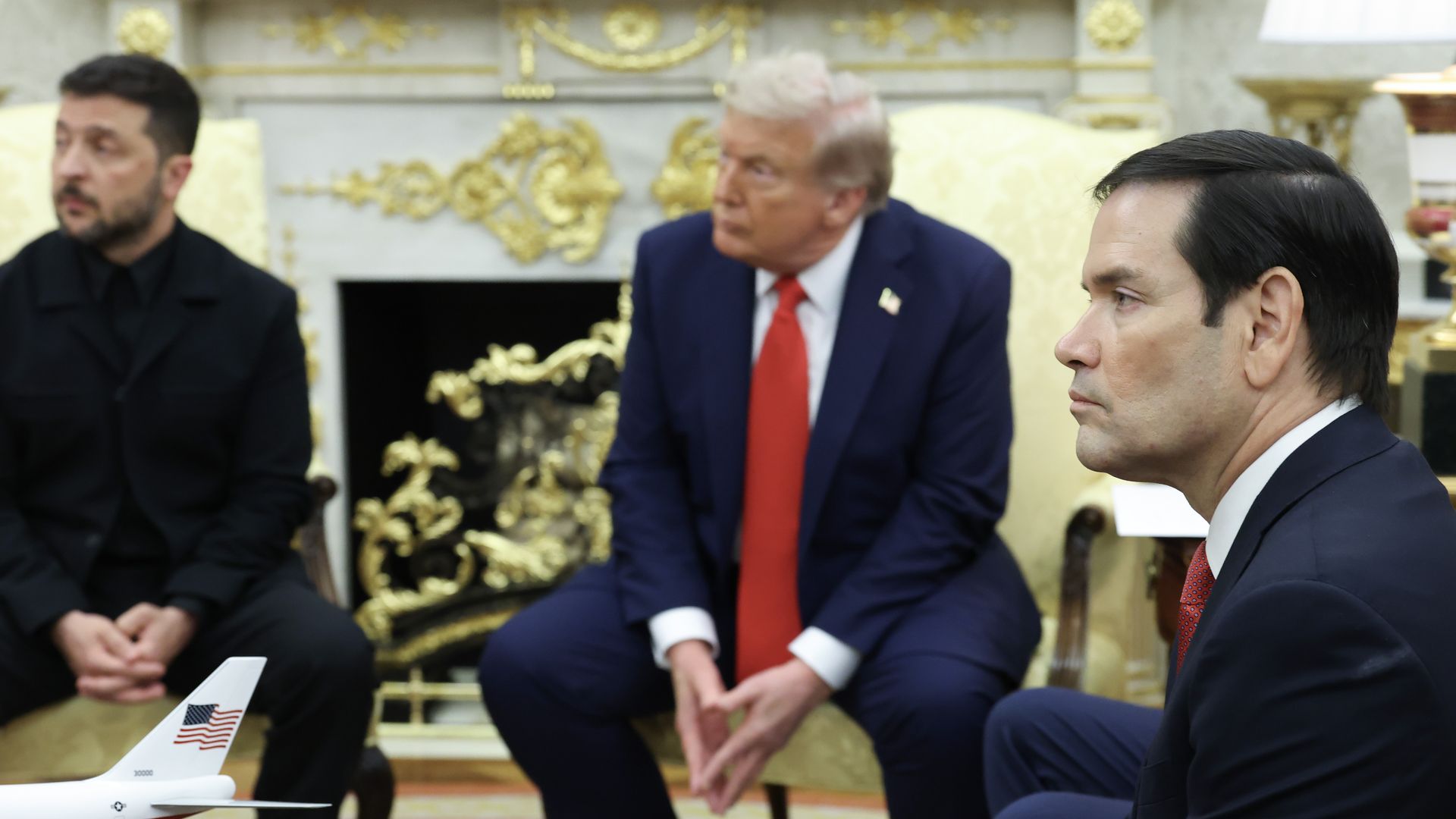 Three men in dark suits seated in an ornate room with a fireplace, decorative gold accents, and a small model airplane with a U.S. flag on the tail in the foreground.