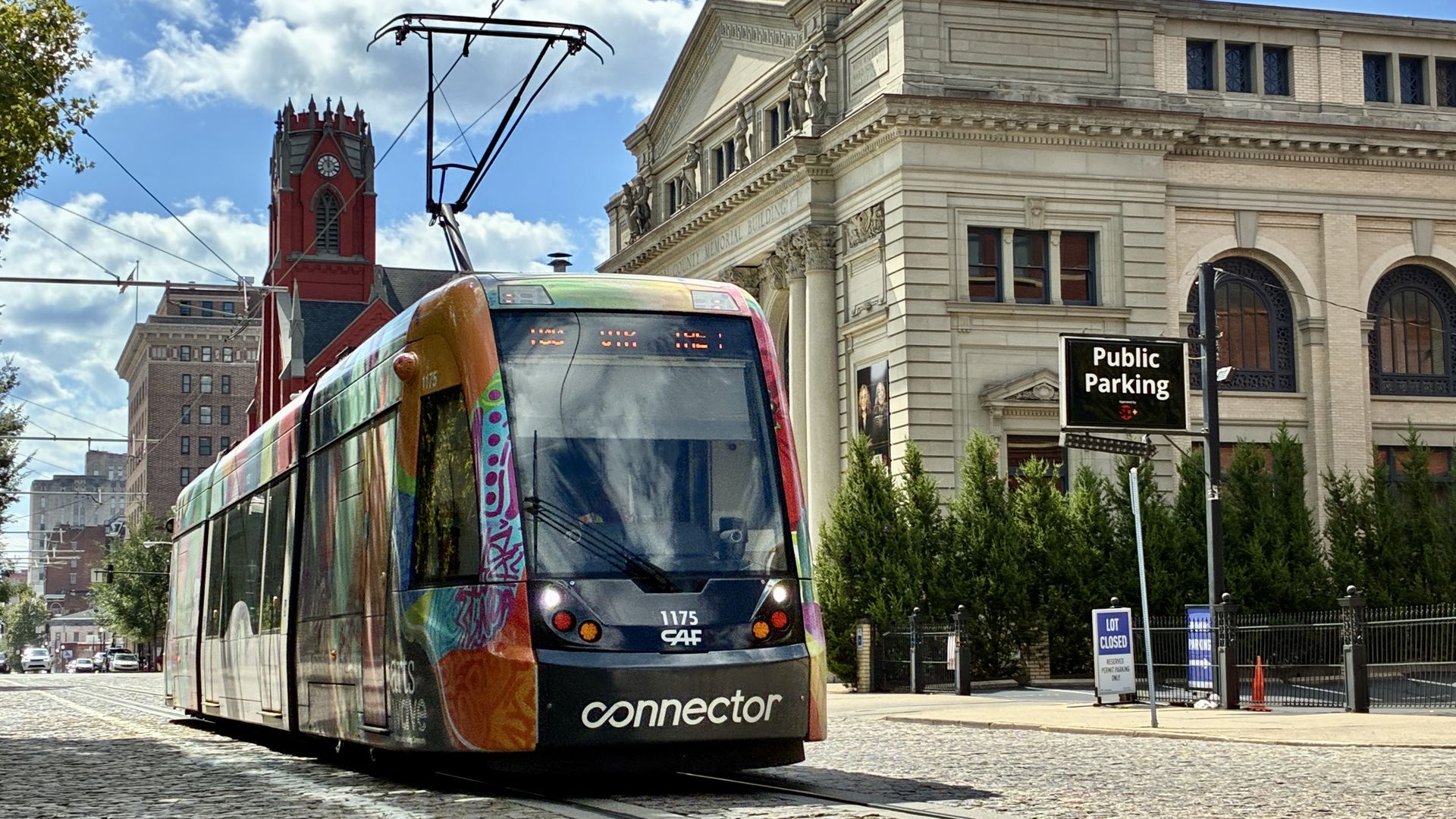 Colorful tram on a cobblestone street in front of a historic cream-colored building with statues; overhead wires crisscross; a red clock tower and blue sky with clouds in the background; Public Parking sign nearby.
