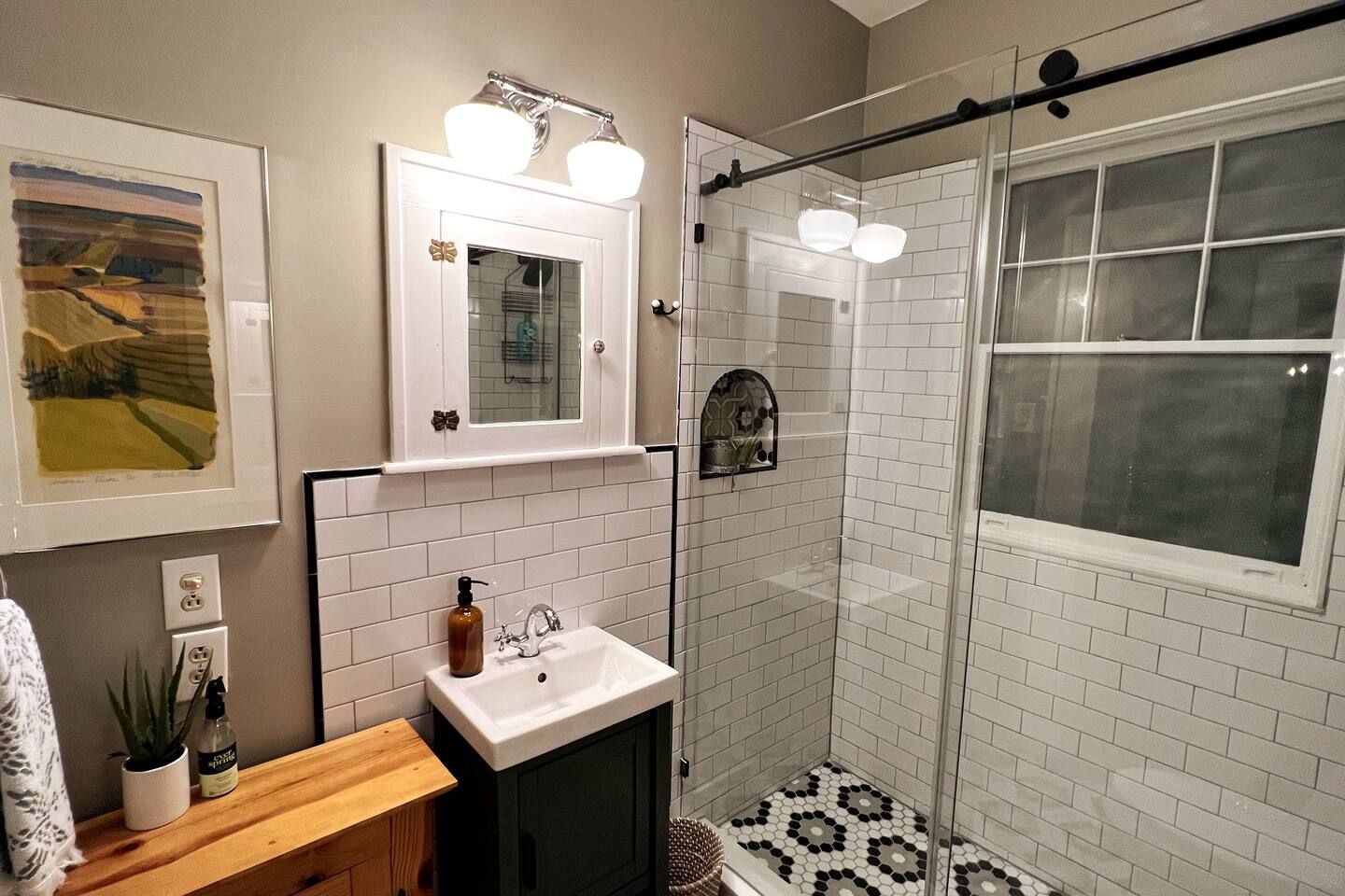 Small bathroom with white subway tile walls and black grout, black and white hexagonal floor tiles, a glass shower door, wooden vanity with white sink, wall-mounted mirror, and beige painted walls.
