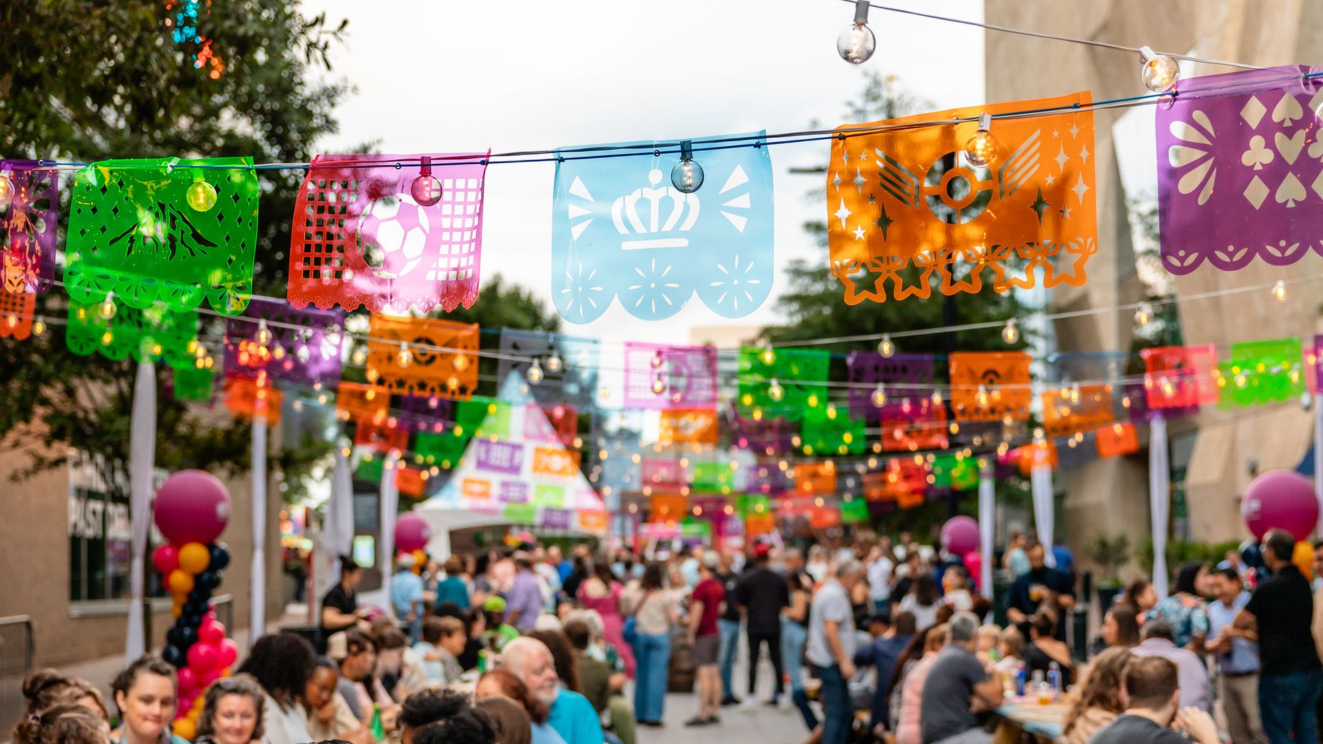 Outdoor community event with many people seated at picnic tables, colorful Mexican papel picado banners and string lights hanging above, festive atmosphere.
