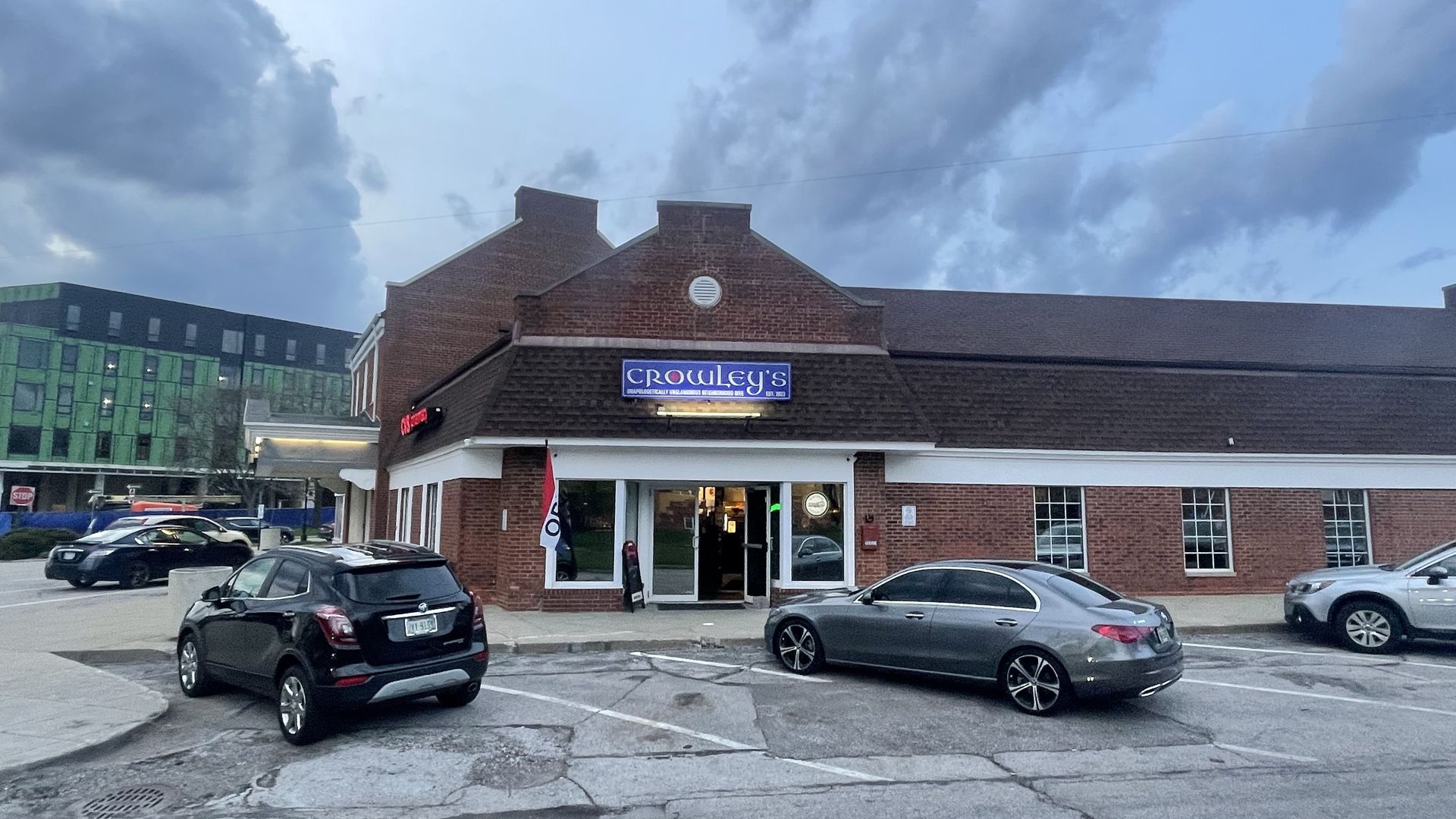 Brick storefront with a blue Crowley’s sign, white trim, and glass doors. Several cars parked in front; a gray-blue sky with clouds; a green construction building visible in the background.