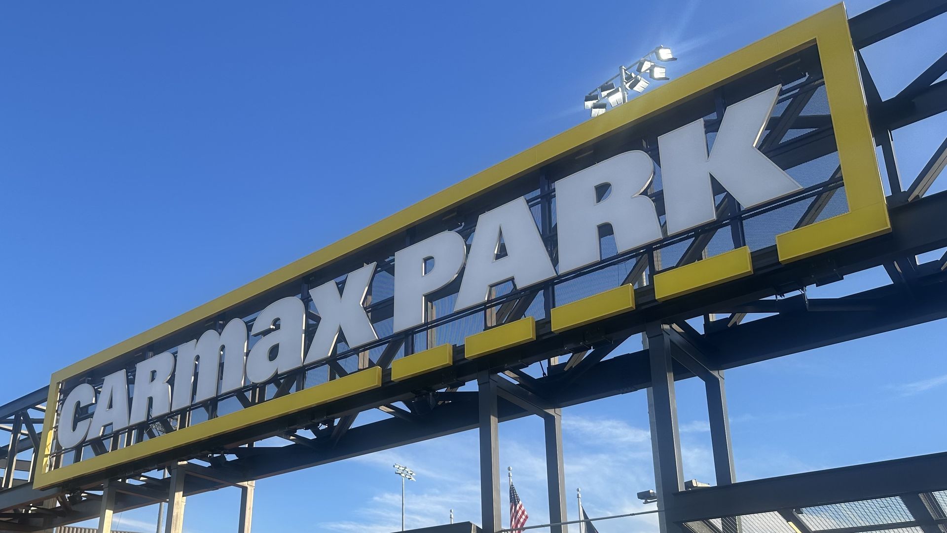 A large entrance sign reading "CARMAXPARK" in white letters framed by a yellow border atop a metal framework, with a clear blue sky and people near the gated entrance below.