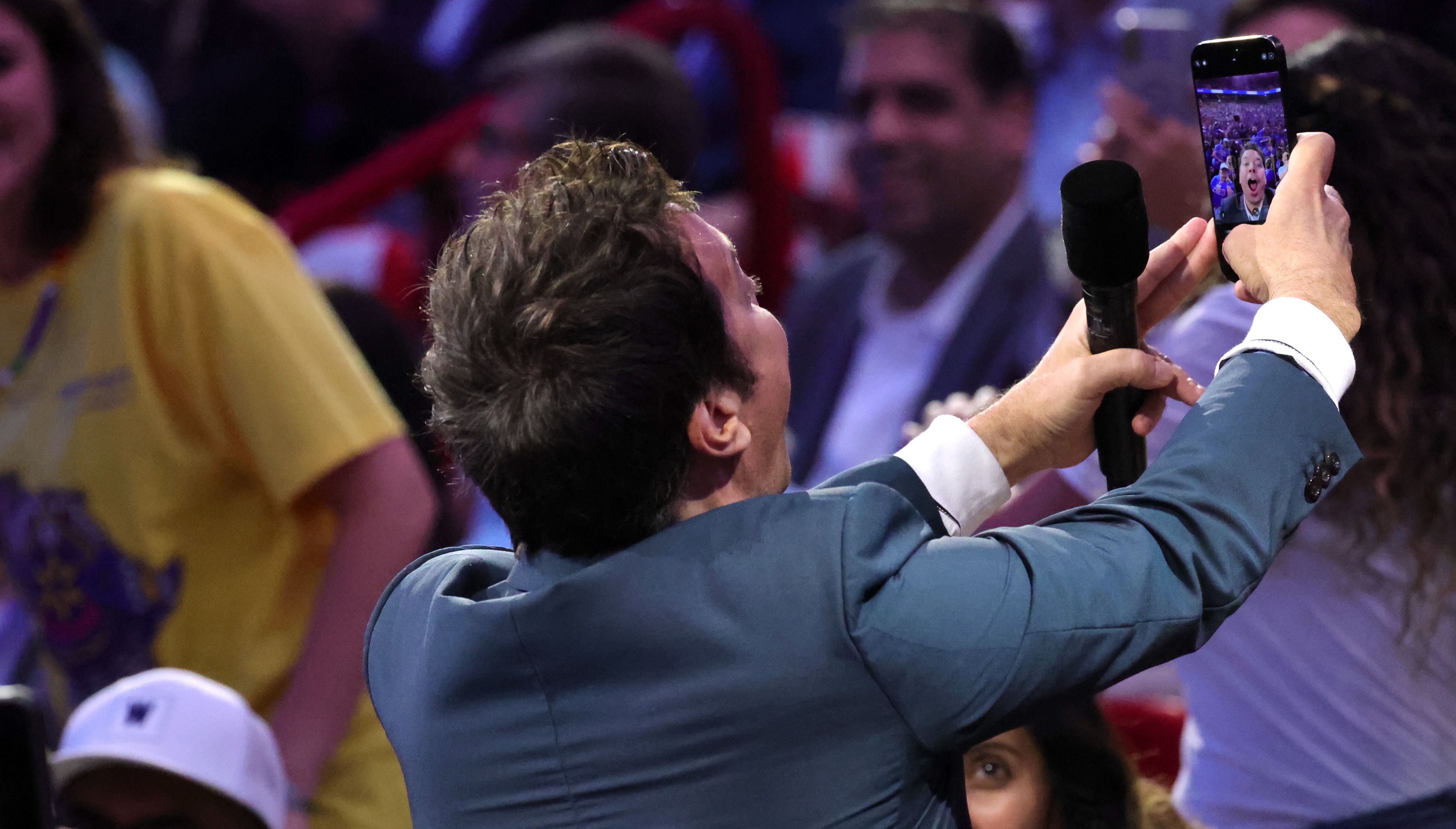 Jimmy Fallon in a a suit holds mic and takes a selfie with crowd behind him, his excited face visible on his phone screen.