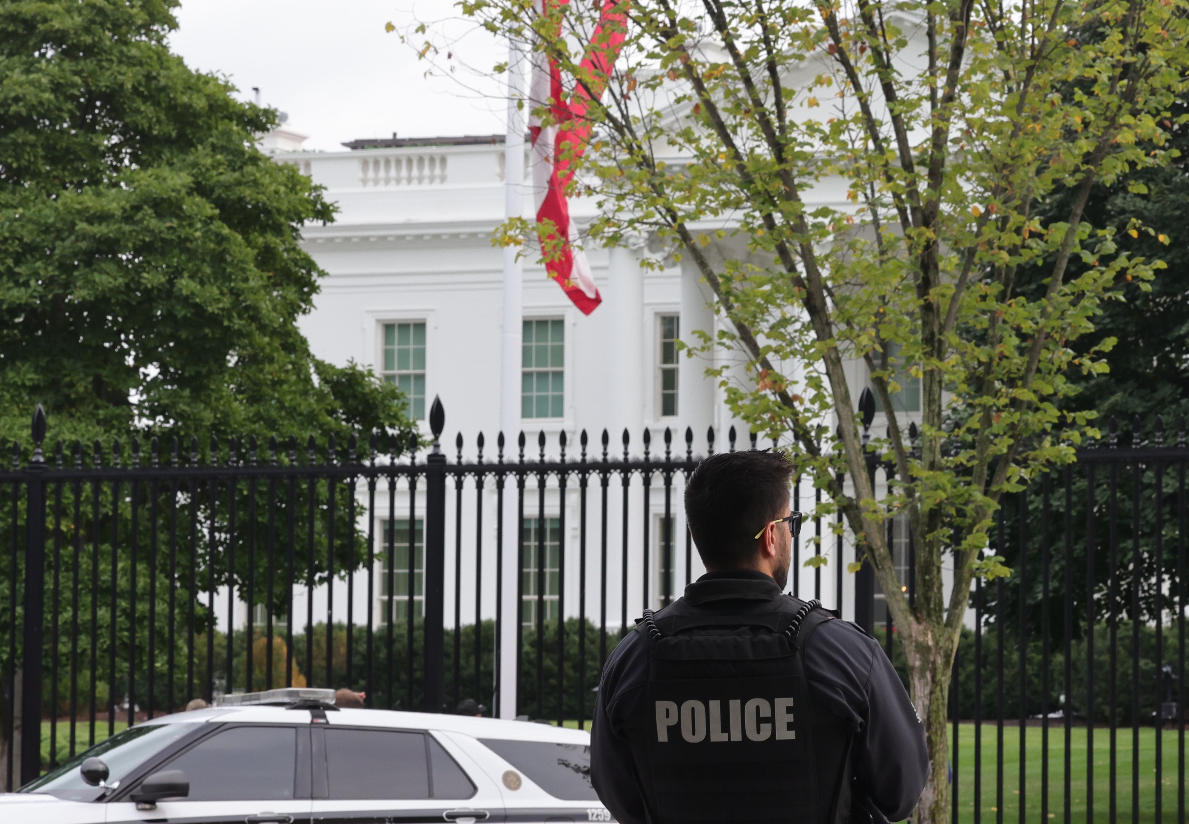 A police officer in black vest stands near a black iron fence, with a white police car parked nearby and the White House partially visible behind trees and the U.S. flag. 