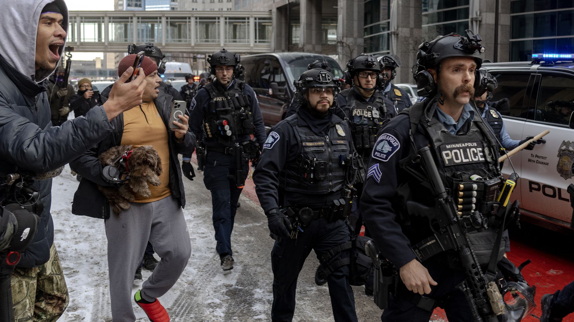 Armored police in tactical gear stand on a snowy street as civilians protest; a shouting man in a gray hoodie holds a phone while a dog sits with another man, blue police lights in the background.