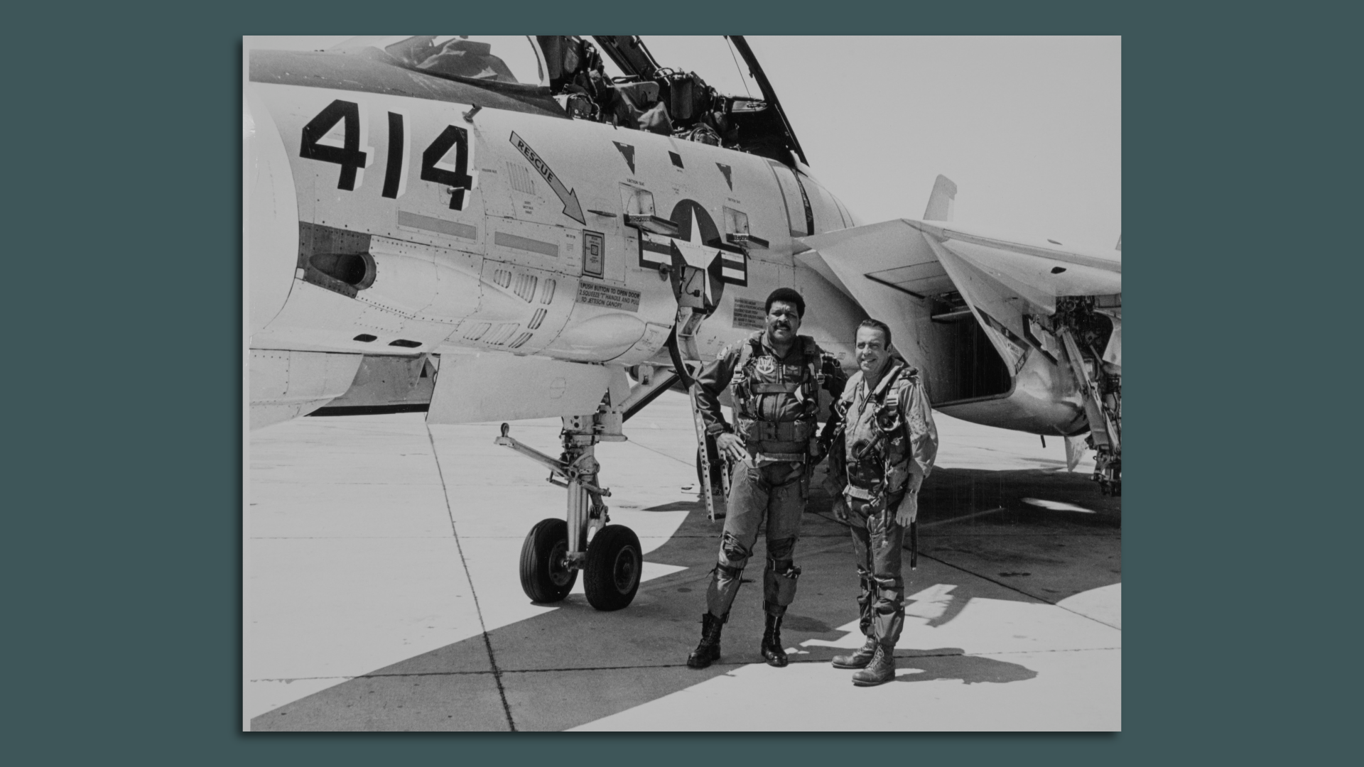 A black and white photo of two military officers in flight gear stand in front of a fighter jet. 