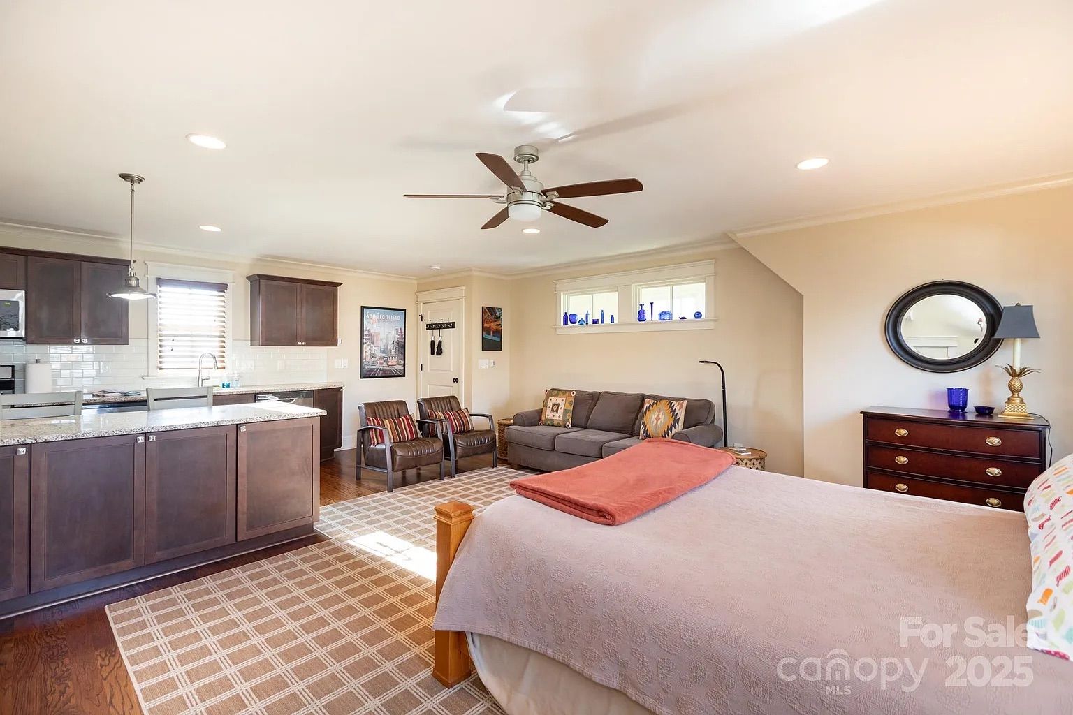 Open-concept room with a bed, two brown leather chairs, gray couch, kitchen with dark cabinets, ceiling fan, and beige plaid rug. Light beige walls with small windows and decor.