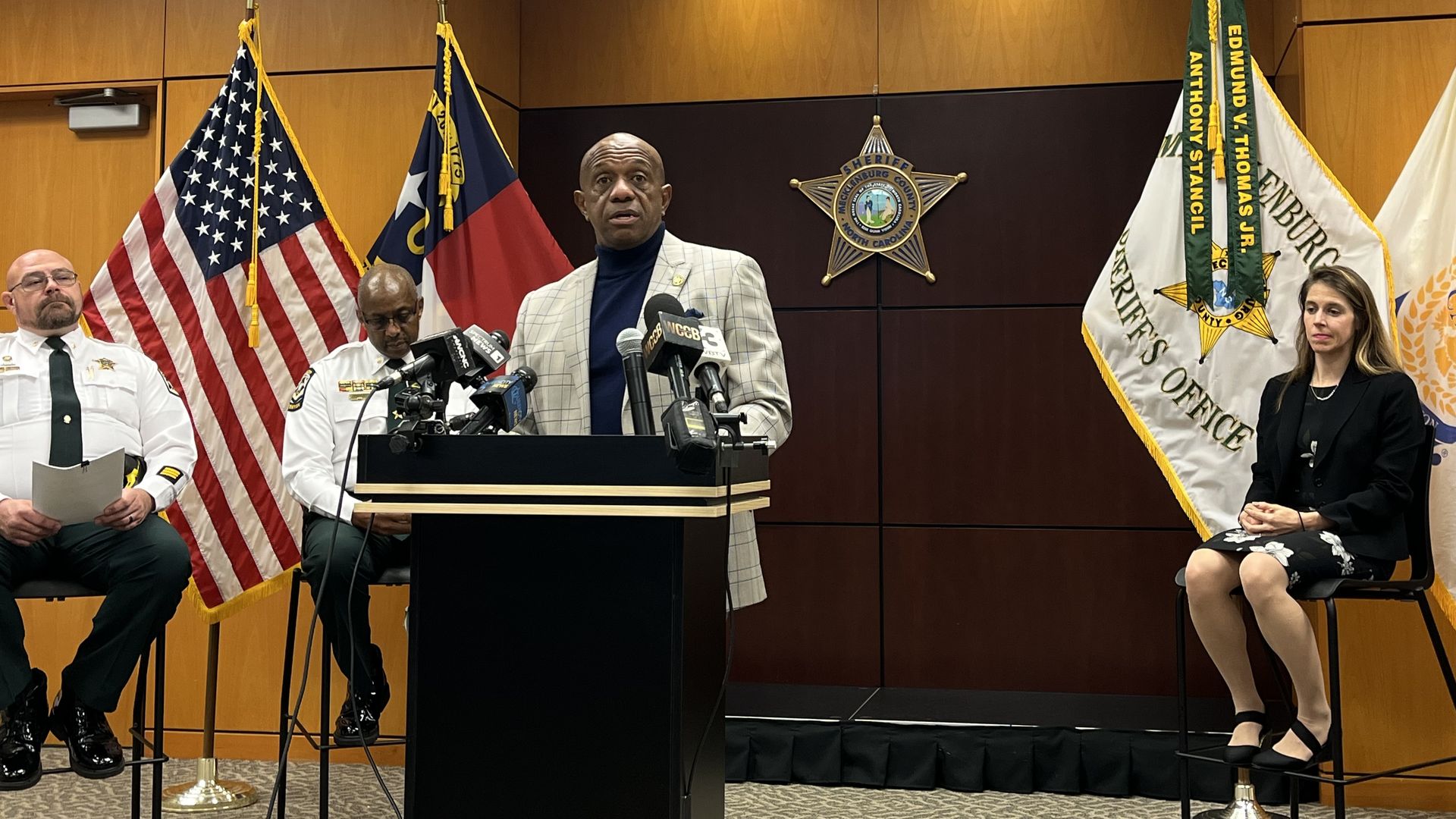 A man in a checkered jacket and navy turtleneck speaks at a podium with microphones in a sheriff's office. Two men in white uniforms and a woman in black sit nearby. Flags and badges behind.