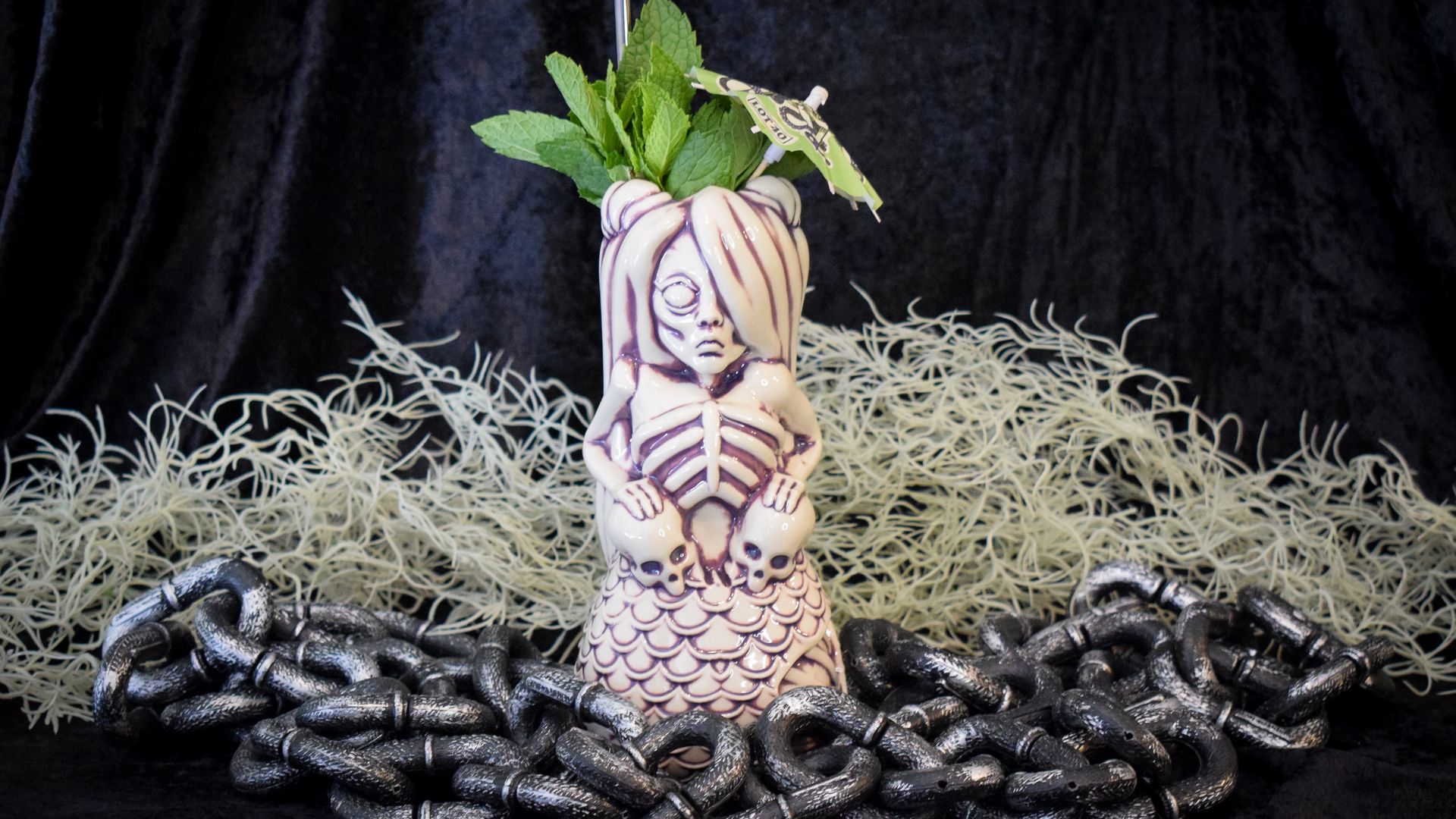 A Tiki glass sits on a black backdrop surrounded by Halloween-themed props like chains and hay.