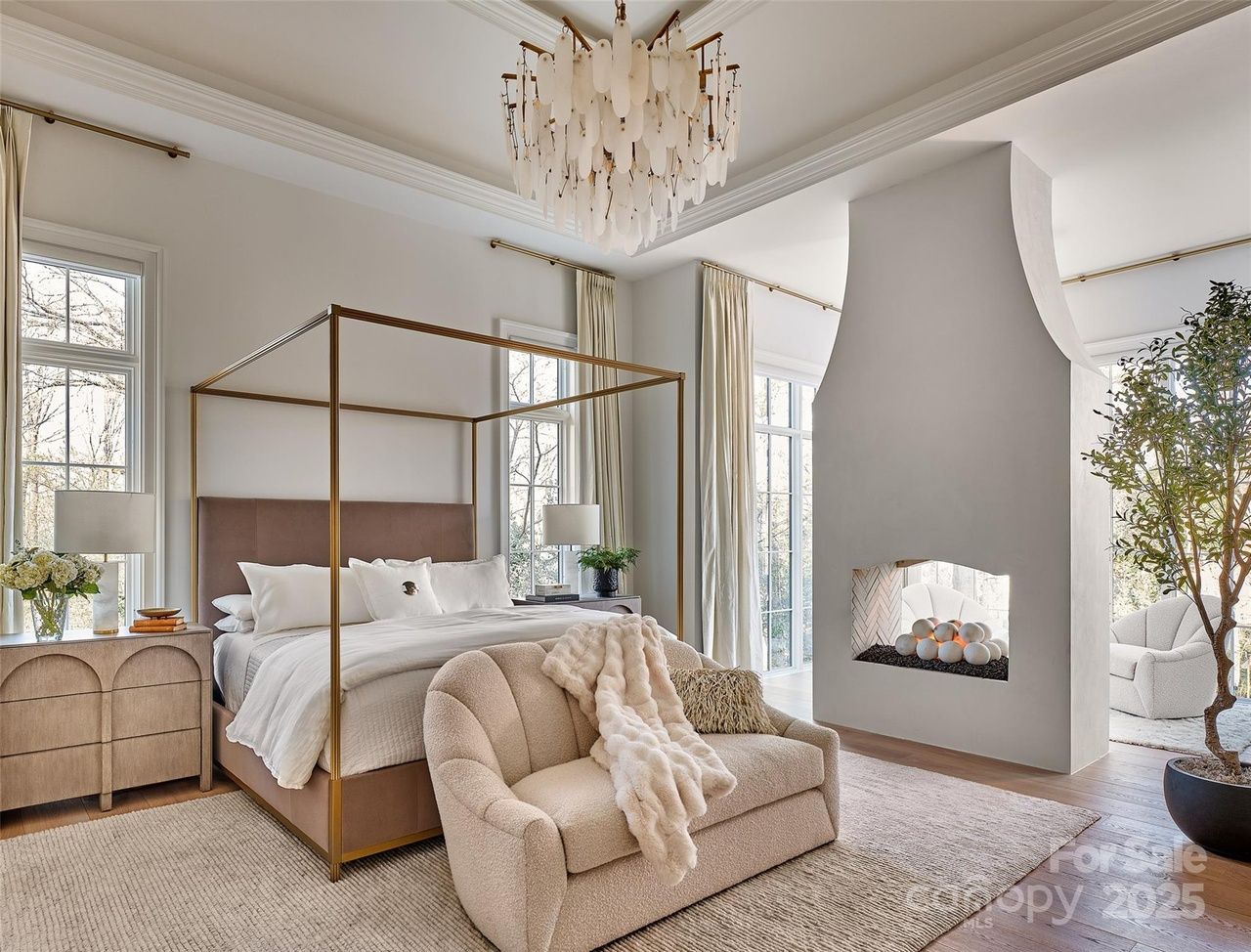 Elegant bedroom with large canopy bed, white linens, beige loveseat with fur throw, modern white fireplace with spherical decor, and tall windows with cream curtains.