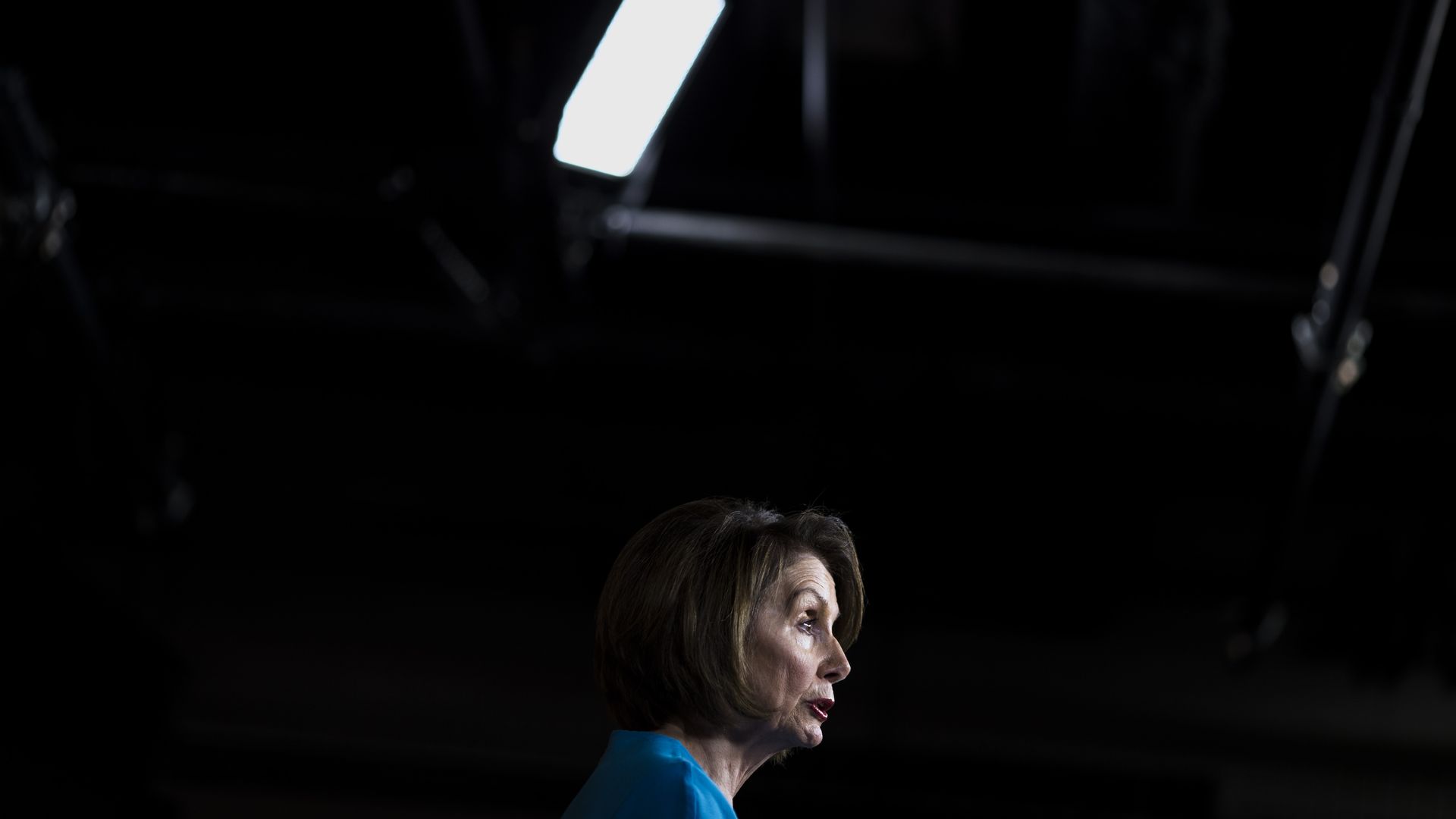 In this image, Nancy Pelosi stands and speaks under a spotlight at a press conference.