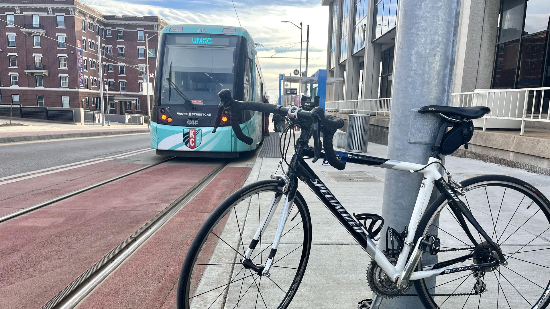 A black and white Specialized road bike locked to a pole on a city sidewalk next to a blue and white RideKC streetcar with UMKC destination displayed, set against urban buildings and a cloudy sky.