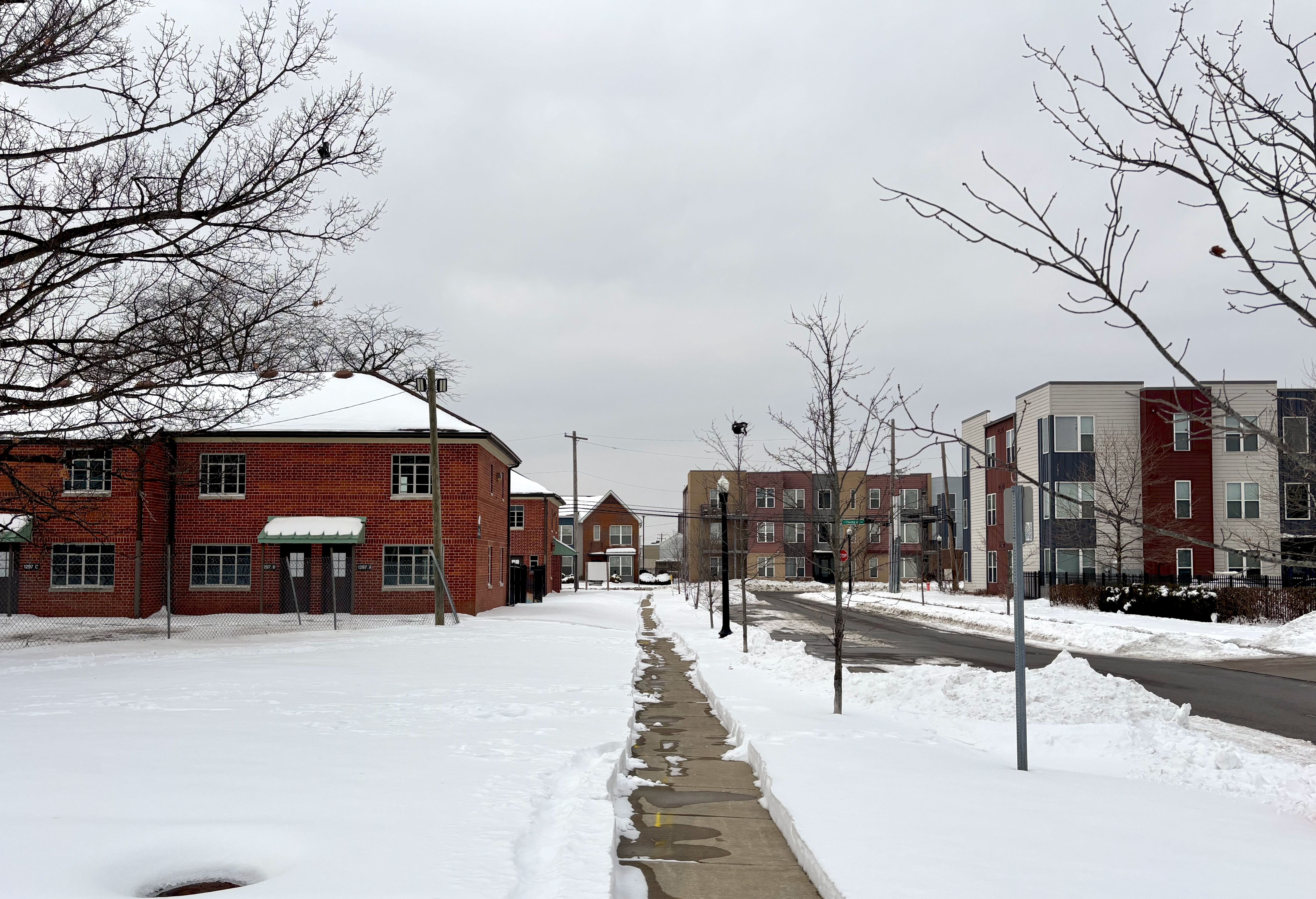 Snow-covered sidewalk between bare trees and residential buildings, with a historic red brick Poindexter Village building ont he left and and modern multi-story homes on the right, under an overcast gray sky in winter.