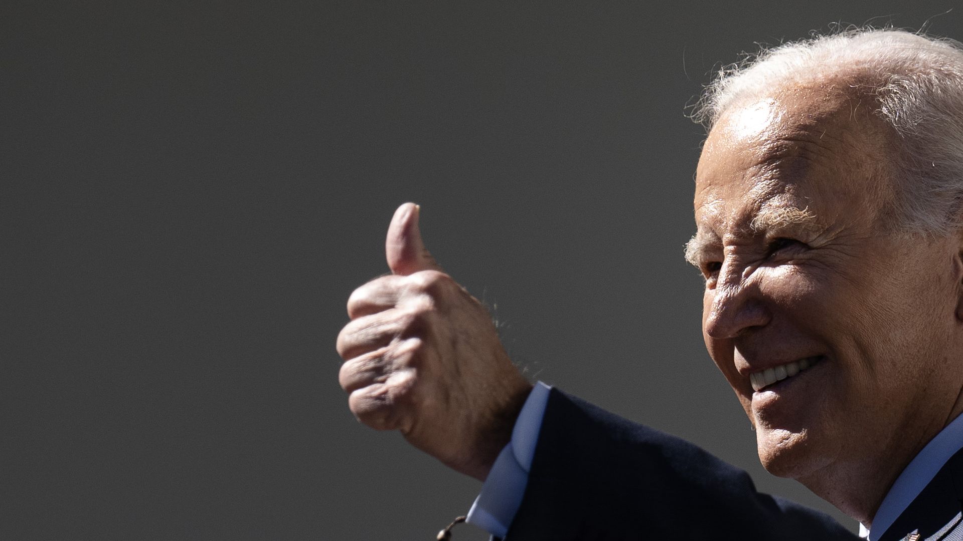 President Joe Biden smiles and squints into the sun, offering a thumbs up sign to someone off camera.