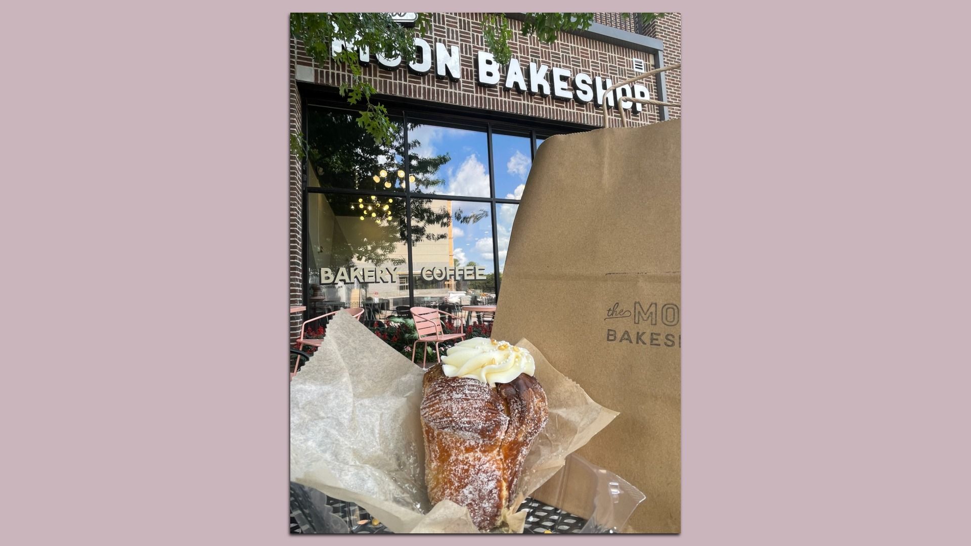 Pastry with white cream on parchment paper in front of a bakery with outdoor pink chairs, large windows, and a brown takeout bag in daylight.