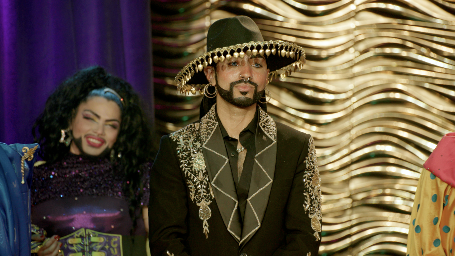 Center: a man in a black, heavily embroidered suit and a wide hat with gold fringe, beard and hoop earrings. A joyful woman in glittery clothing stands to his left; purple curtain and golden wavy backdrop behind.