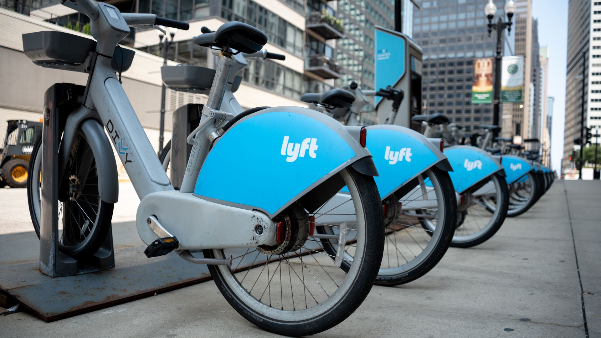 Line of light-blue Lyft bikes docked at a city bike-share station; foreground shows a bike frame, while tall buildings, a lamppost, and banners loom in the background.