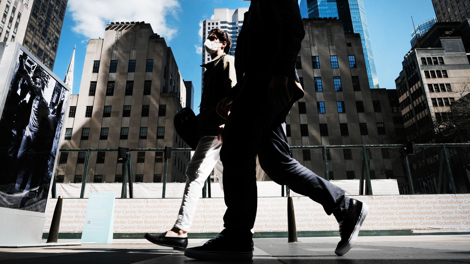 Two men walk on a sidewalk in front of tall buildings, shot from the street level.