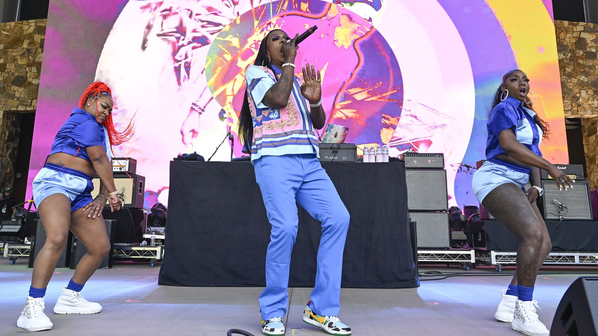 Big Freedia sings into a microphone while two dancers perform to either side of her.