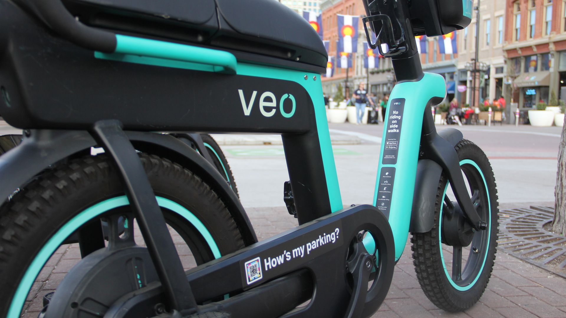 Close-up of a black and teal Veo electric bike with thick tires parked on a city sidewalk with buildings and Colorado state flags in the background.