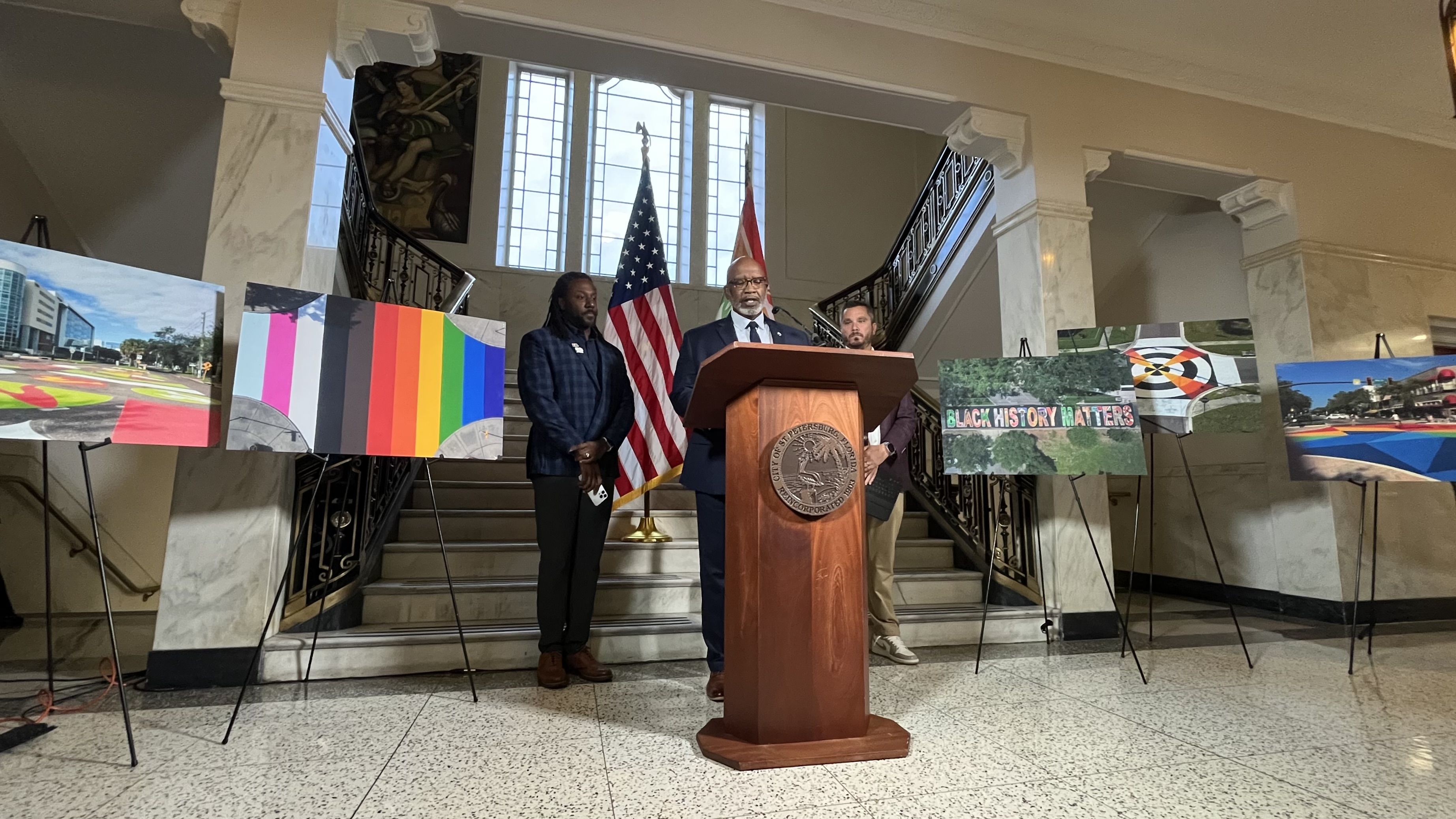 Three men stand at a podium in a marble hall with a US flag and another flag behind them. Posters on easels show colorful street art, including LGBTQ+ pride and Black History themes.