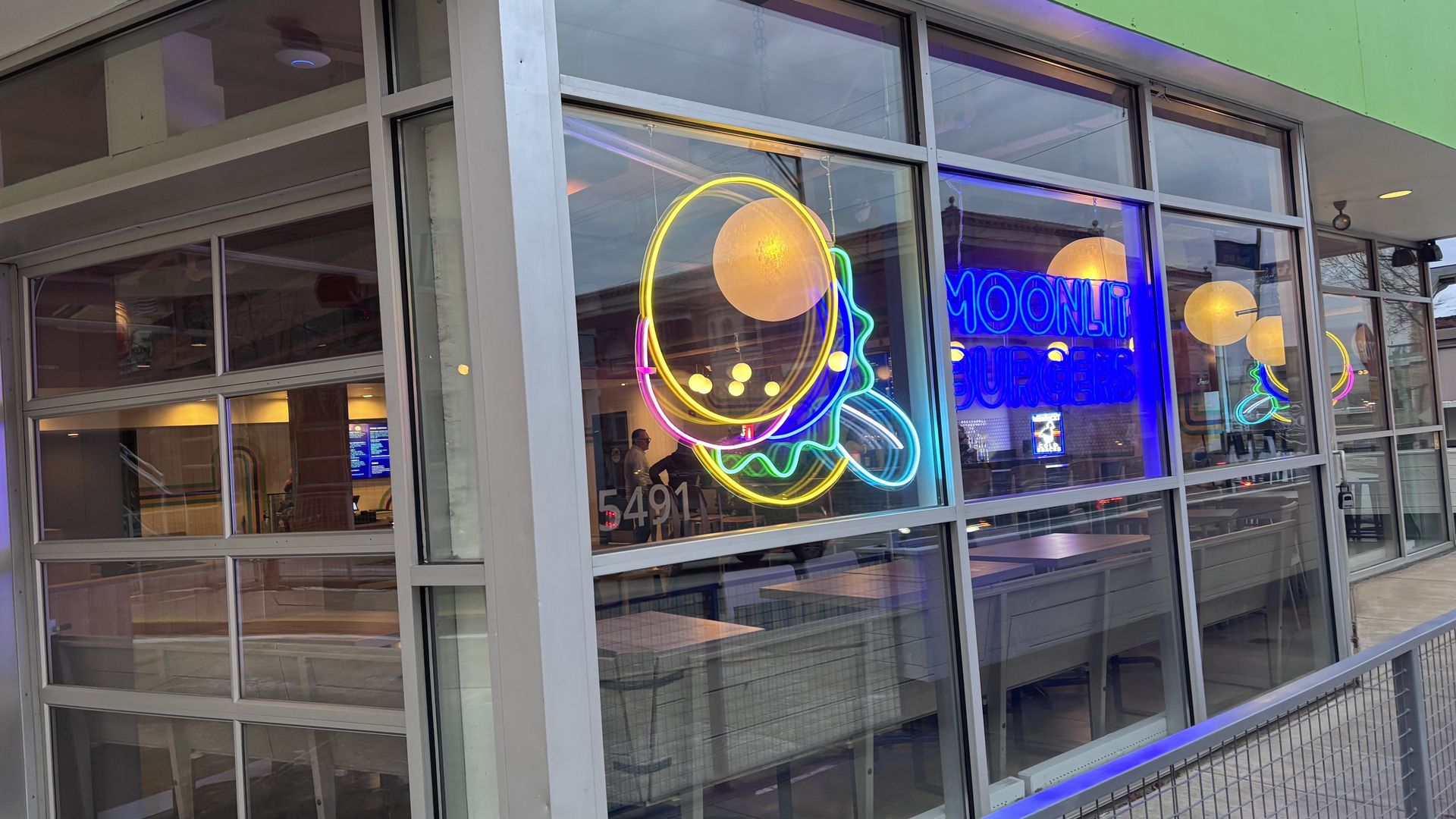 Exterior view of a restaurant with large windows displaying colorful neon signs of a burger and the text "Moonlit Burgers", with tables and hanging round lights inside.