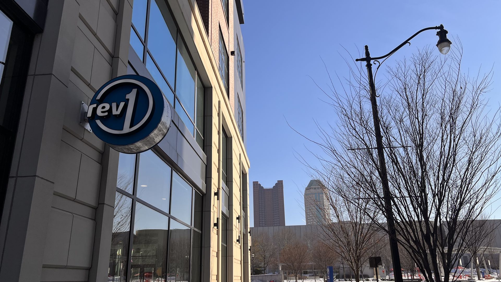Sidewalk with snow beside a beige building featuring a blue circular "rev1" sign, bare trees, a streetlamp, parked car, and distant tall buildings under a clear blue sky.